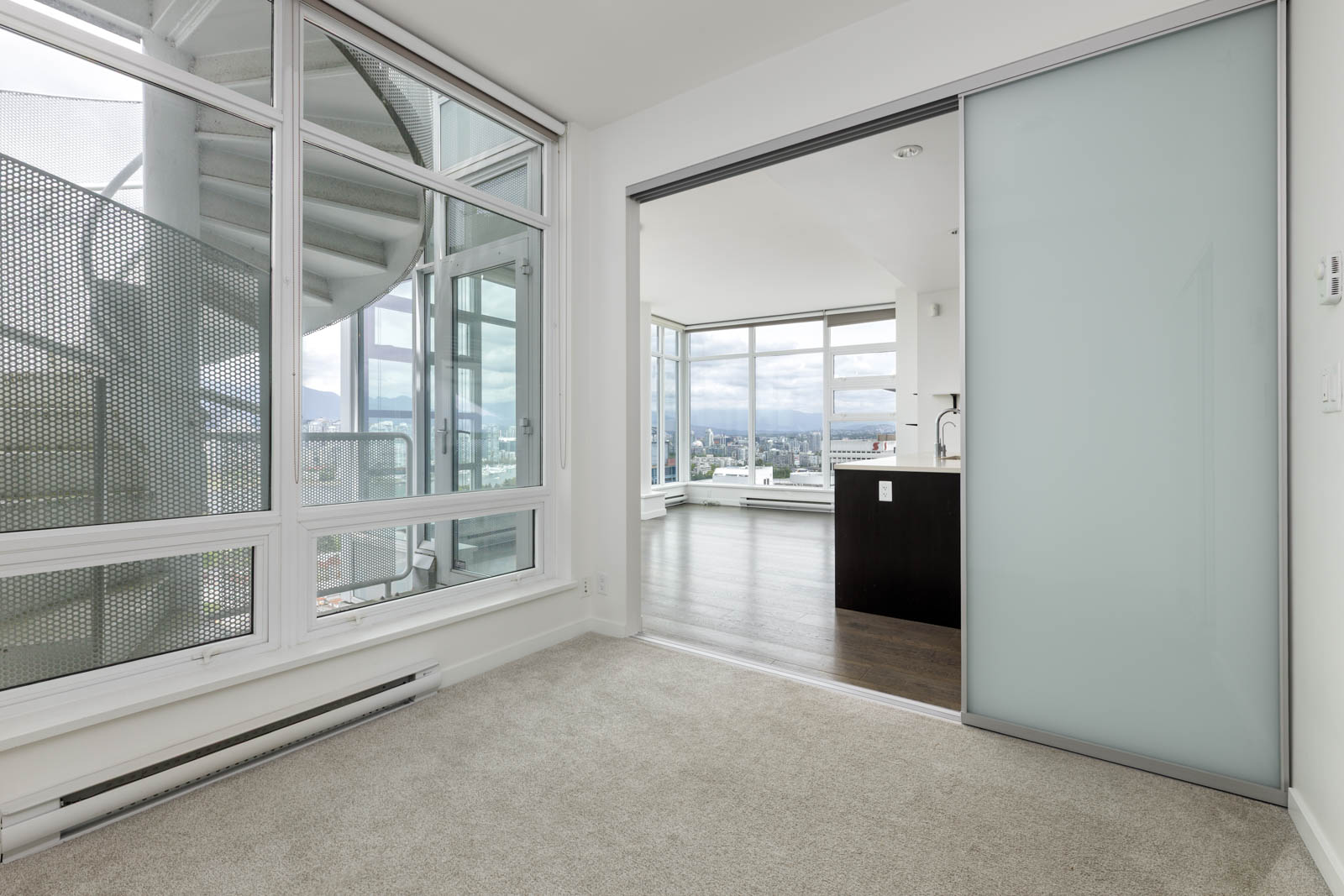 Modern apartment interior with carpeted room, sliding frosted glass door, large windows, balcony spiral staircase, and view of cityscape in the background.
