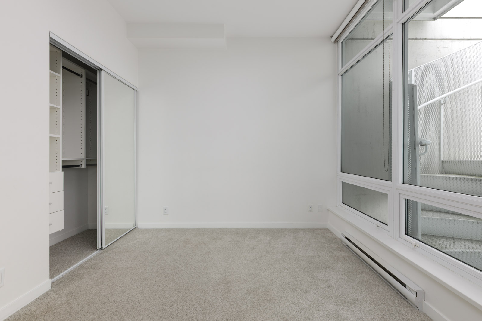 Empty room with beige carpet, white walls, a sliding mirrored closet door on the left, and large windows on the right with an outside view of a staircase.