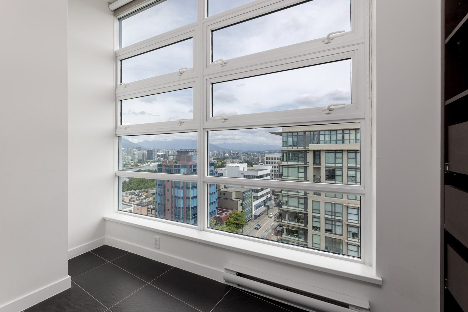 Large windows in a high-rise room offer a view of modern city buildings and a cloudy sky, with black tiled flooring visible in the foreground.