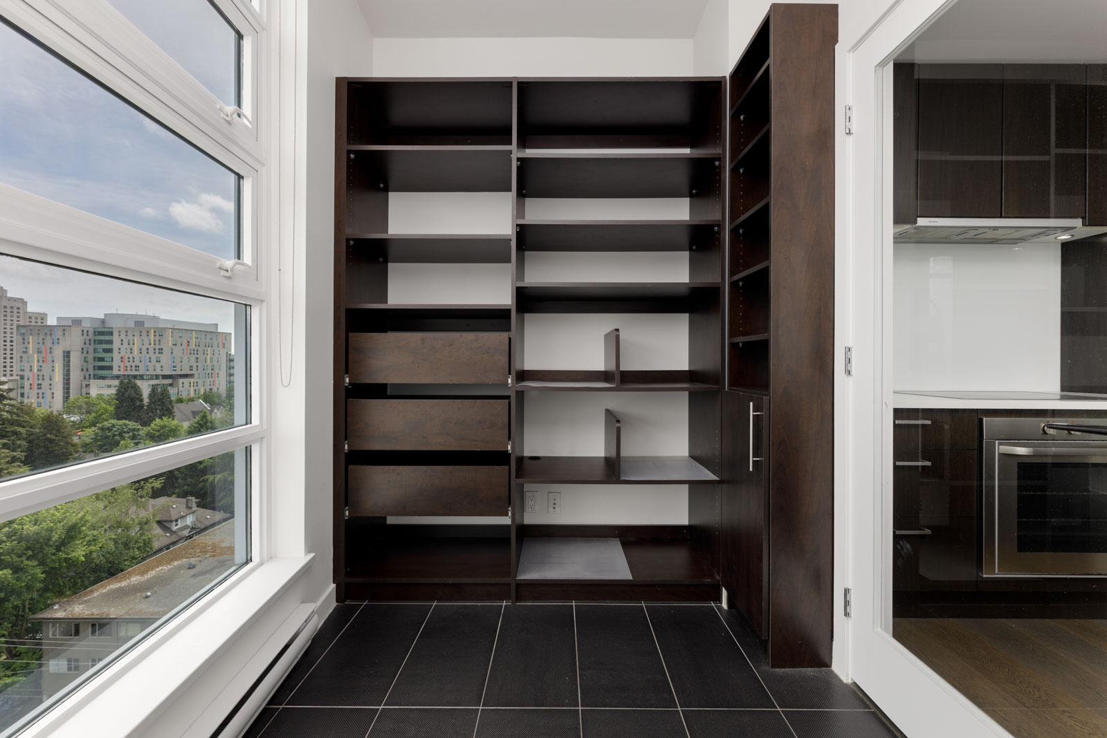 A modern pantry with dark wood shelves, empty and built into a corner, next to large windows with a city view and adjacent to a kitchen.