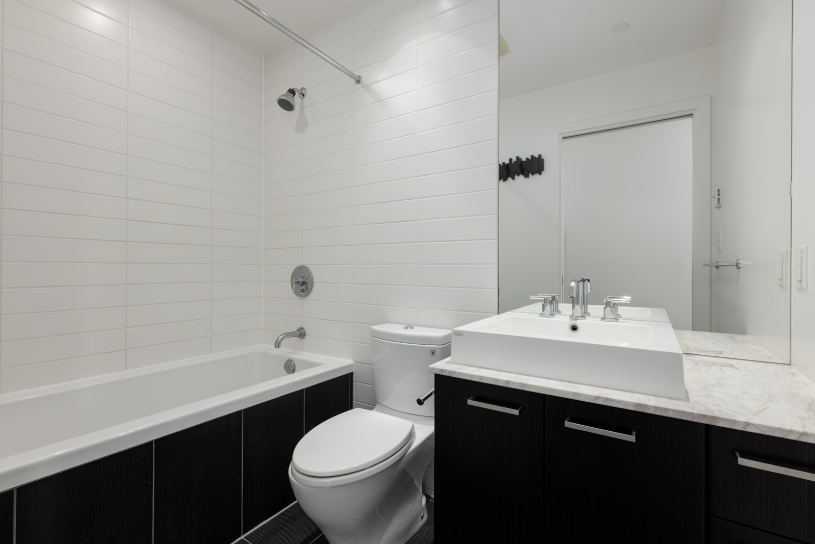 Modern bathroom with white tiled walls, bathtub, toilet, and a sink with a marble countertop and dark vanity, viewed from the doorway.