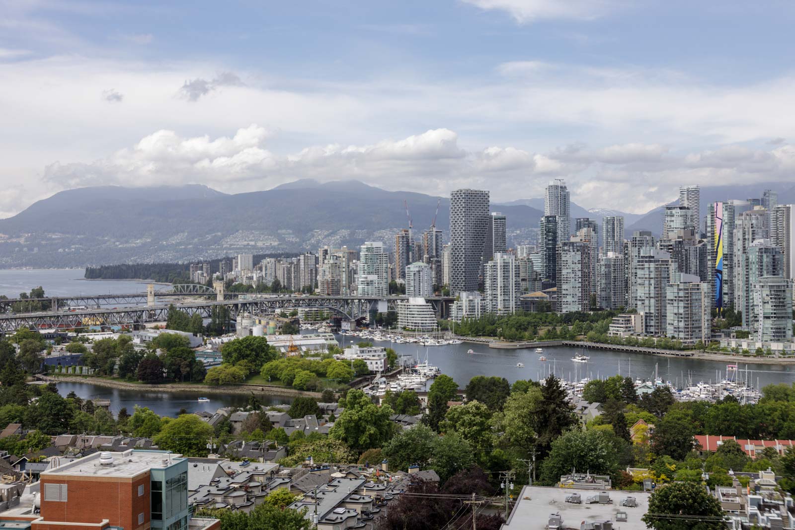 A cityscape of Vancouver, featuring modern high-rise buildings, a river with boats, tree-lined areas, and mountains in the background under a partly cloudy sky.