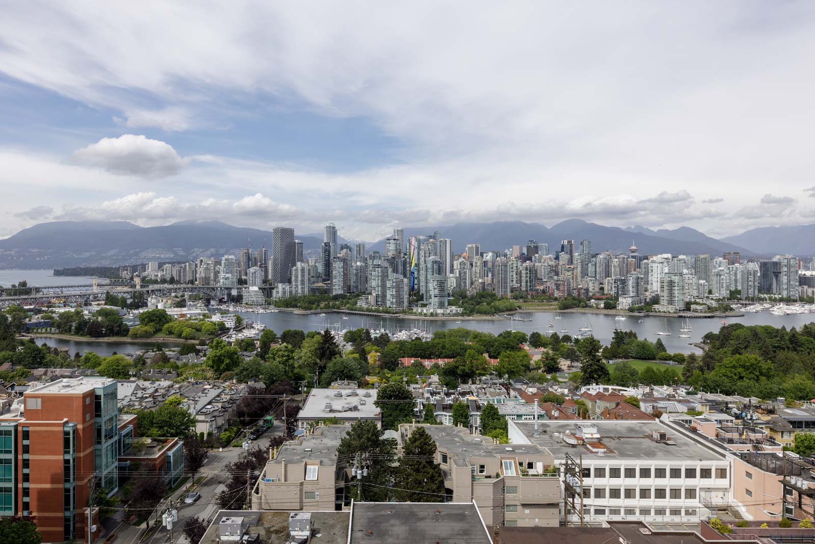 Aerial view of a city skyline with tall buildings, a river, and mountains in the background under a partly cloudy sky.