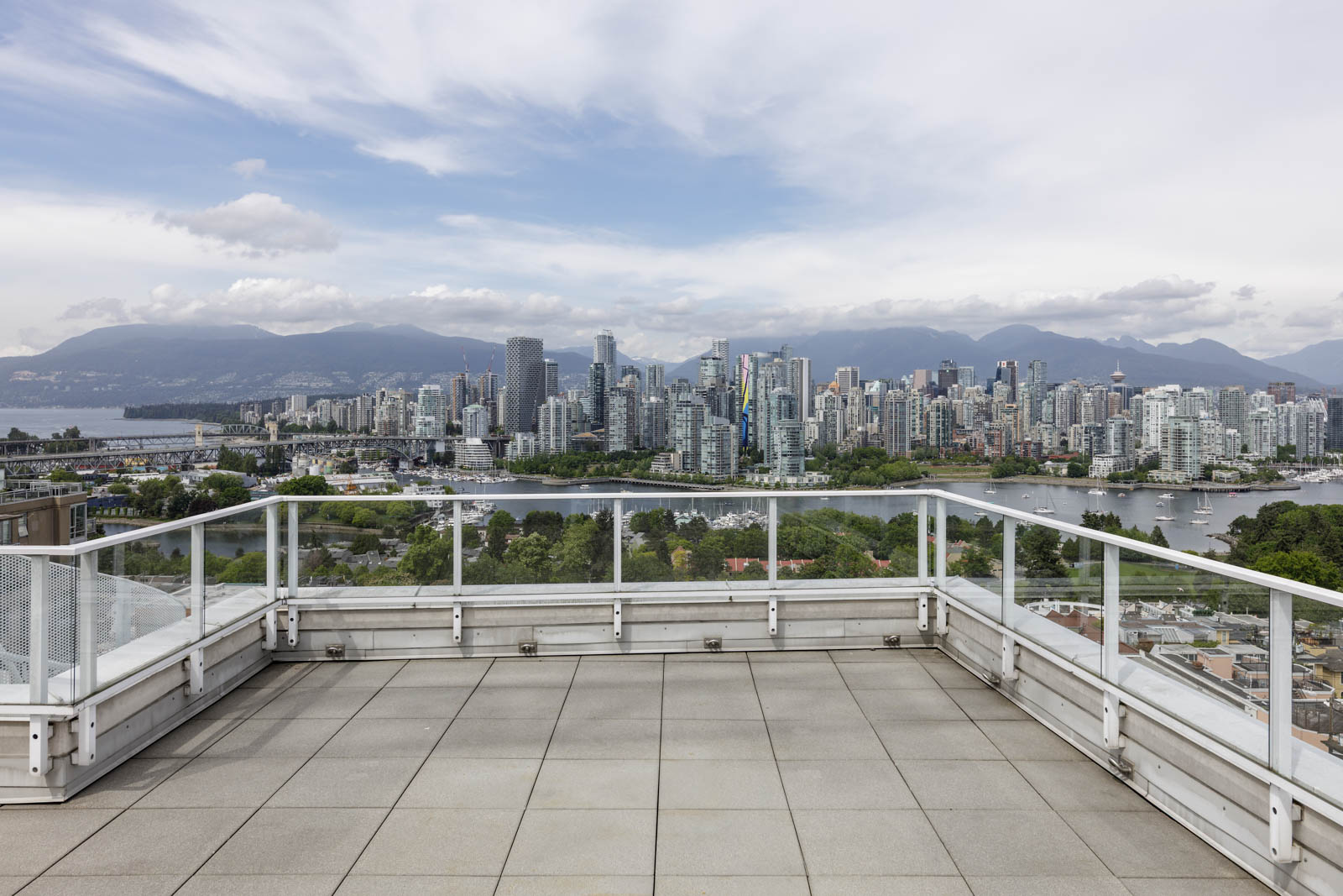 A rooftop terrace with glass railings overlooks a city skyline, featuring high-rise buildings, greenery, and mountains in the background under a cloudy sky.