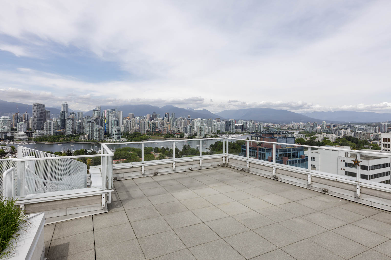 Rooftop terrace with tile flooring and glass railing overlooking a city skyline, river, and distant mountains under a mostly cloudy sky.