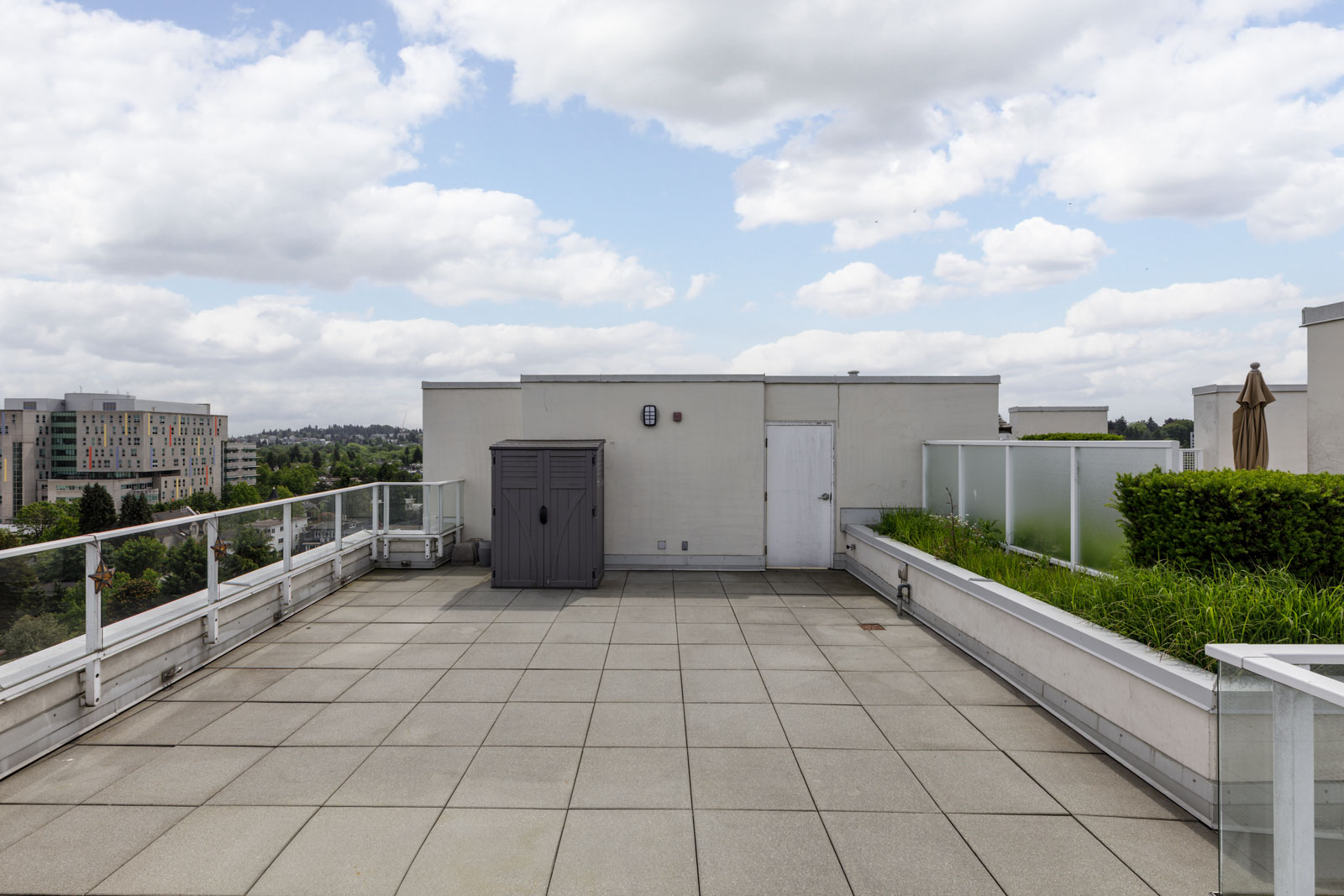 A rooftop patio with tiled flooring, a storage shed, planters with greenery, and glass railings, under a partly cloudy sky.