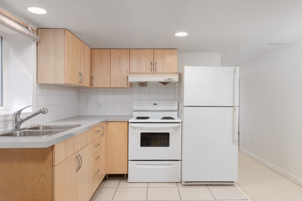 Compact kitchen with light wood cabinets, white electric stove, refrigerator, double sink, tiled floor, and white walls under recessed lighting.