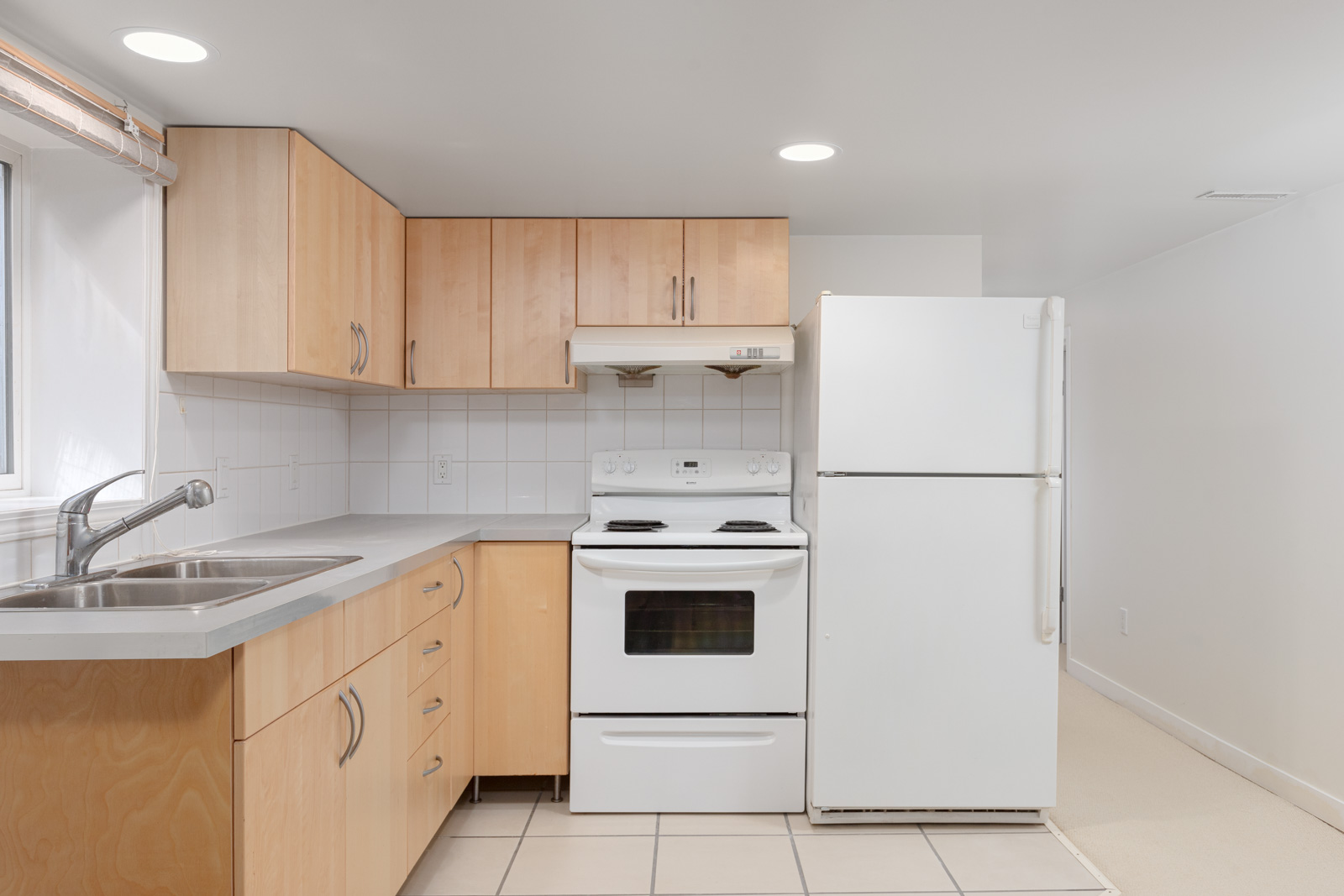 Compact kitchen with light wood cabinets, white electric stove, refrigerator, double sink, tiled floor, and white walls under recessed lighting.