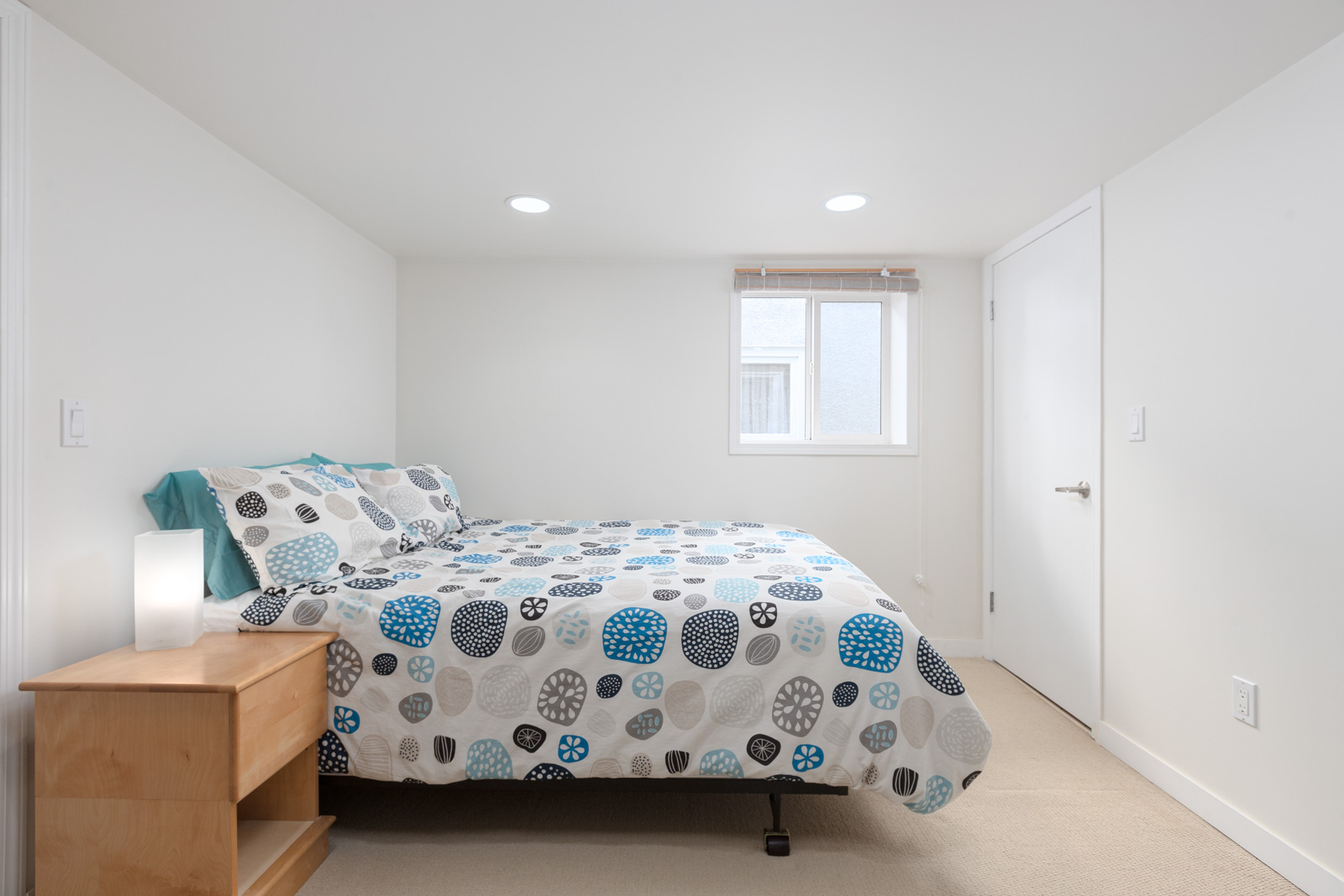 Minimalist bedroom with a double bed, polka dot bedding, wooden nightstand with a lamp, beige carpet, white walls, and a small window letting in natural light.