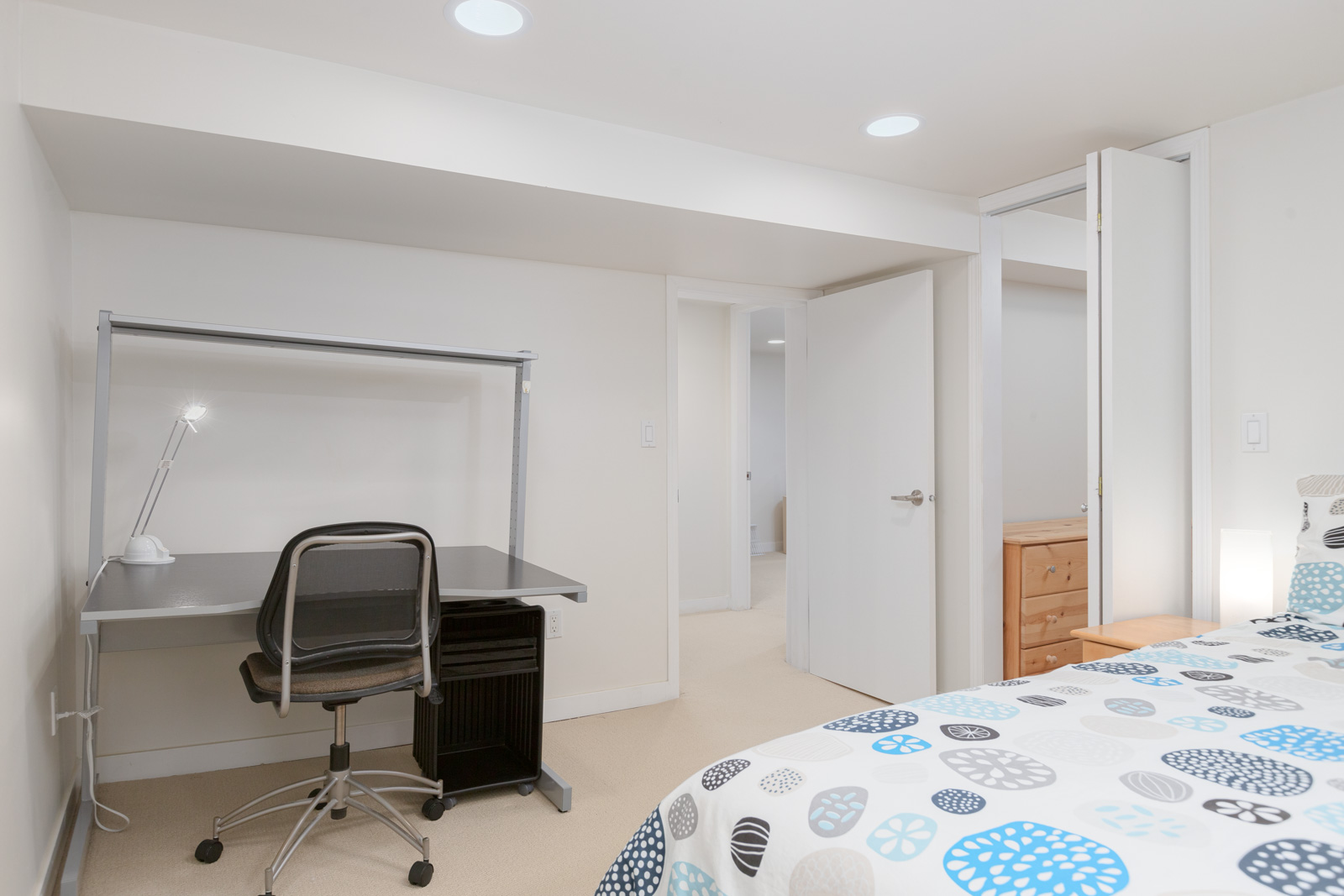 A tidy bedroom with a bed, a black desk and chair, a desk lamp, and an open door leading to a hallway with light-colored walls and carpet.