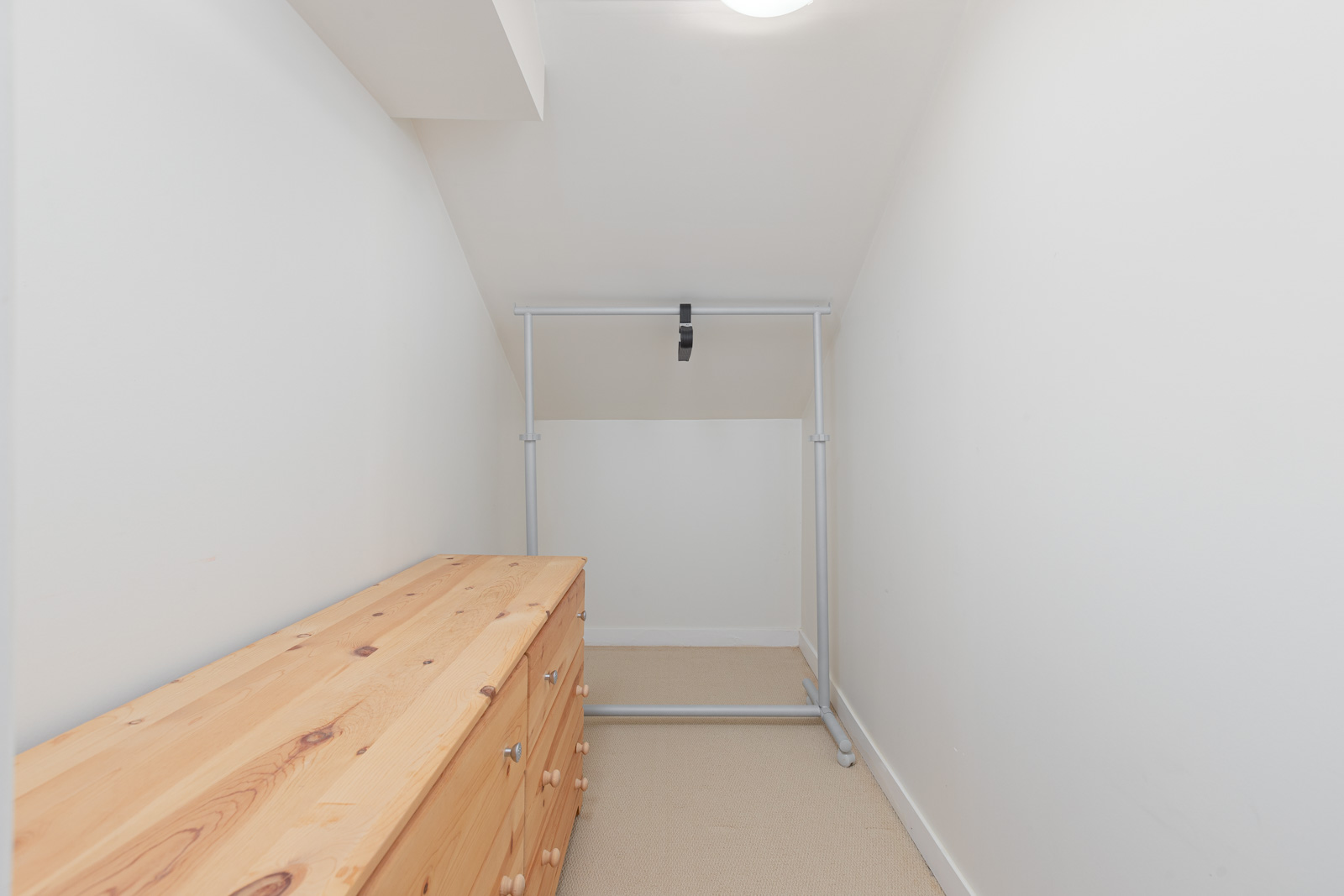 A small, narrow room with white walls, a wooden dresser on the left, and a metal clothing rack at the end under a ceiling light.