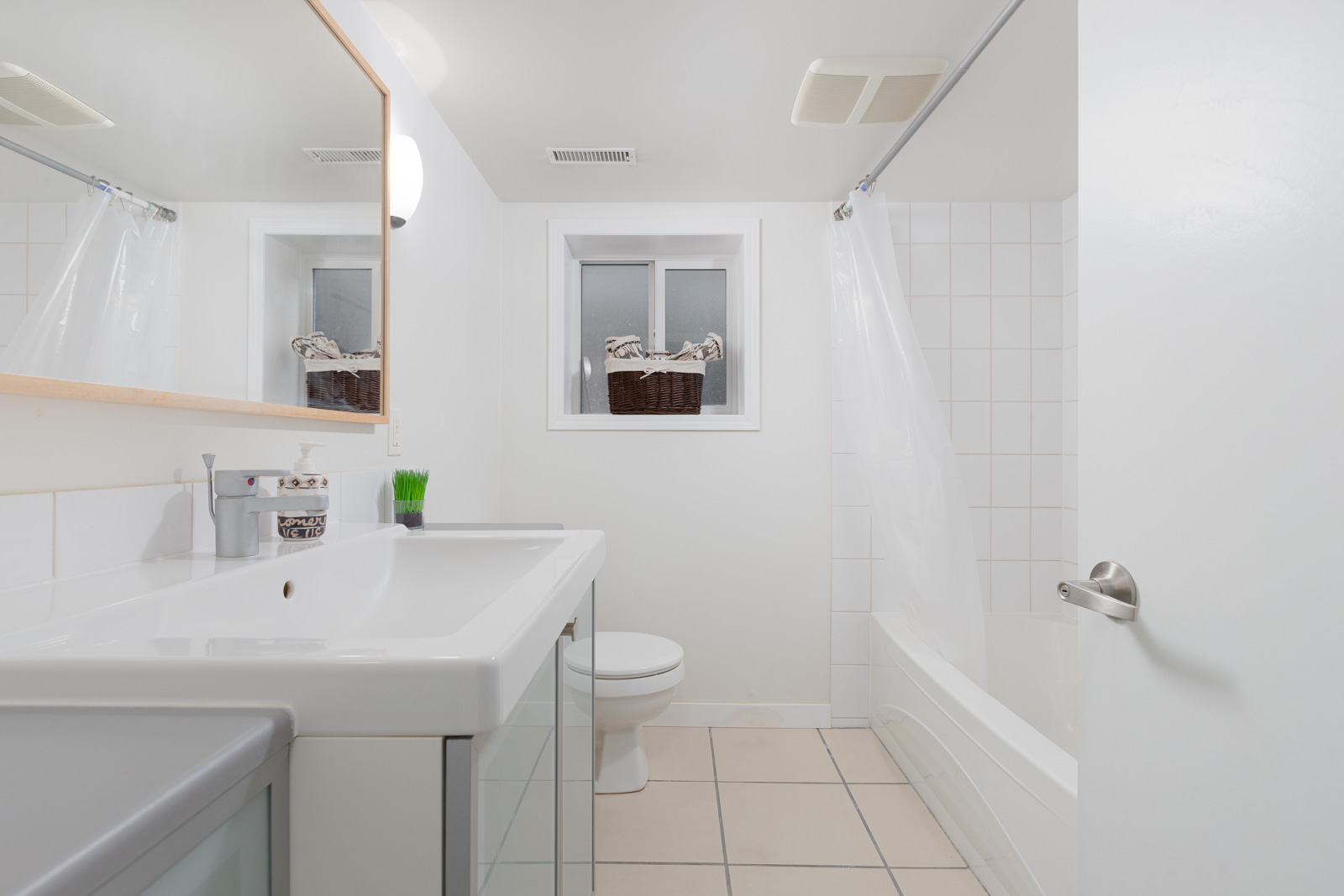 A clean, white bathroom with a sink, toilet, bathtub with a shower curtain, tile flooring, and a window above the toilet. Two brown baskets are on the window ledge.