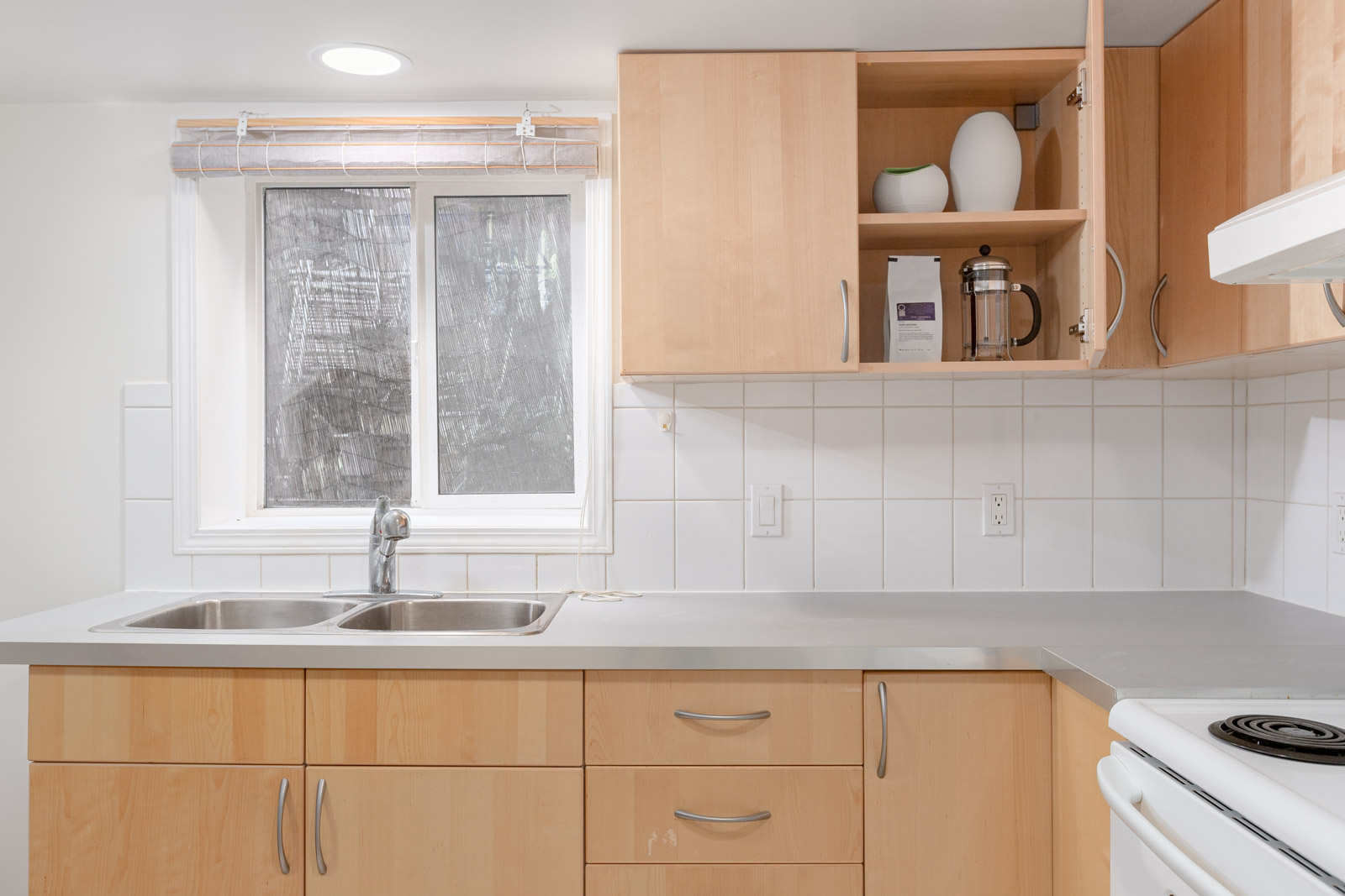 A modern kitchen with light wood cabinets, a double sink, white tile backsplash, and open shelving displaying decor and a French press.