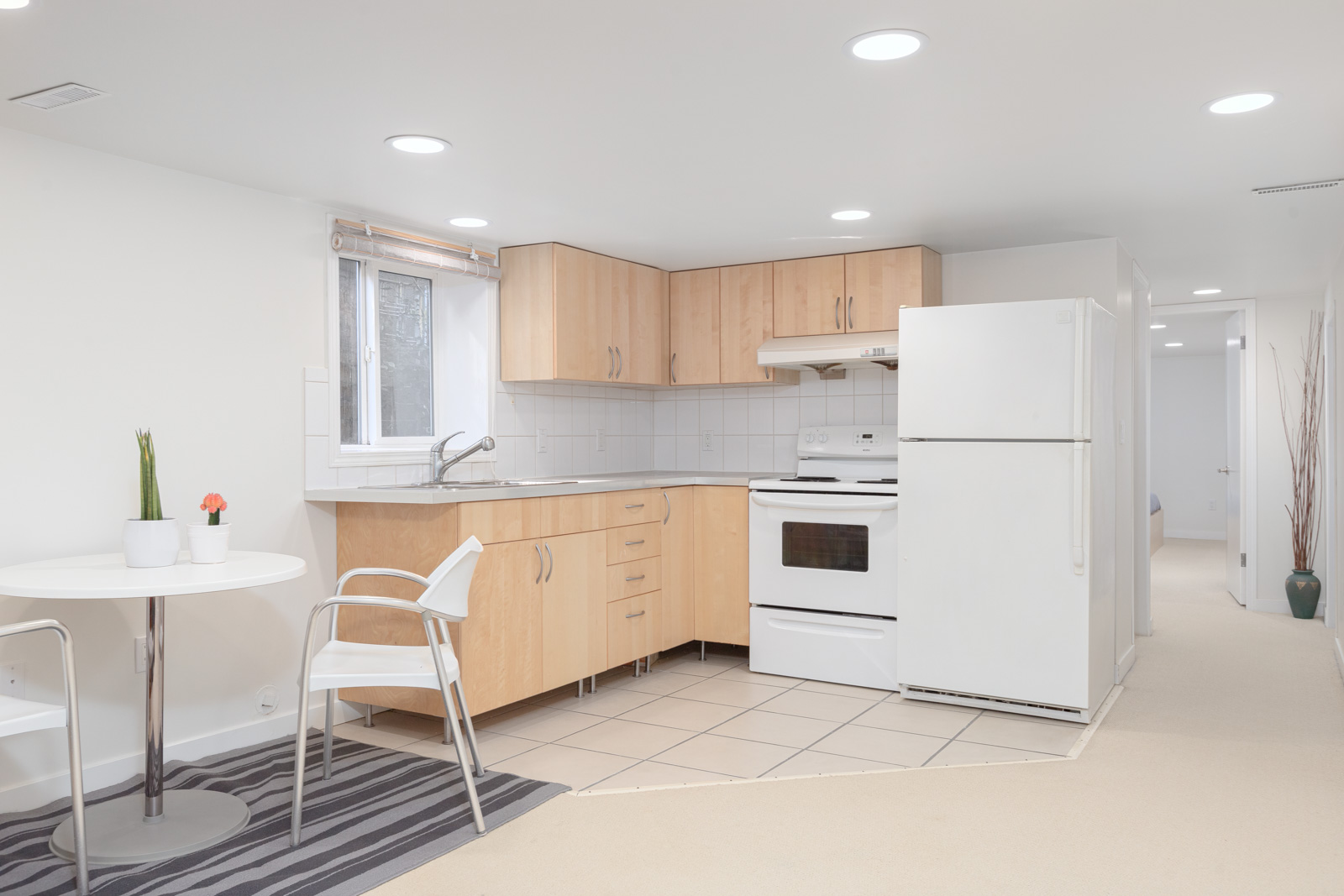 Compact kitchen and dining area with light wood cabinets, white appliances, a small round table with two chairs, and minimal decor in a bright, clean space.