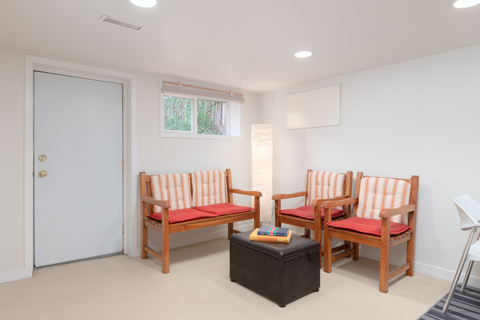 A small waiting room with two wooden benches with striped cushions, a floor lamp, a window, a closed door, and a black ottoman with books on top.