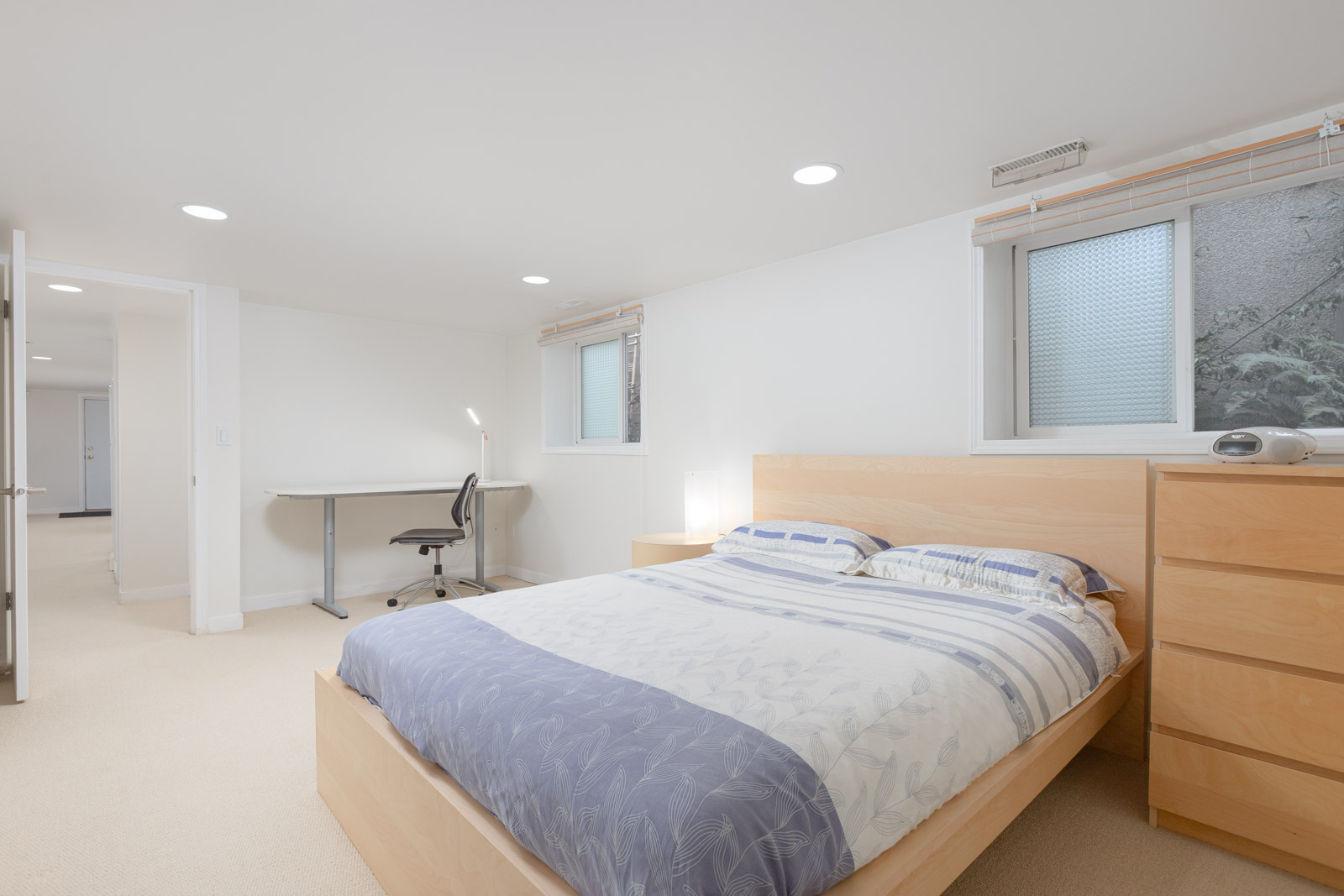 A modern bedroom with a wooden bed, matching dresser and nightstand, a desk with a chair, and two windows, all in a bright, minimalist setting.