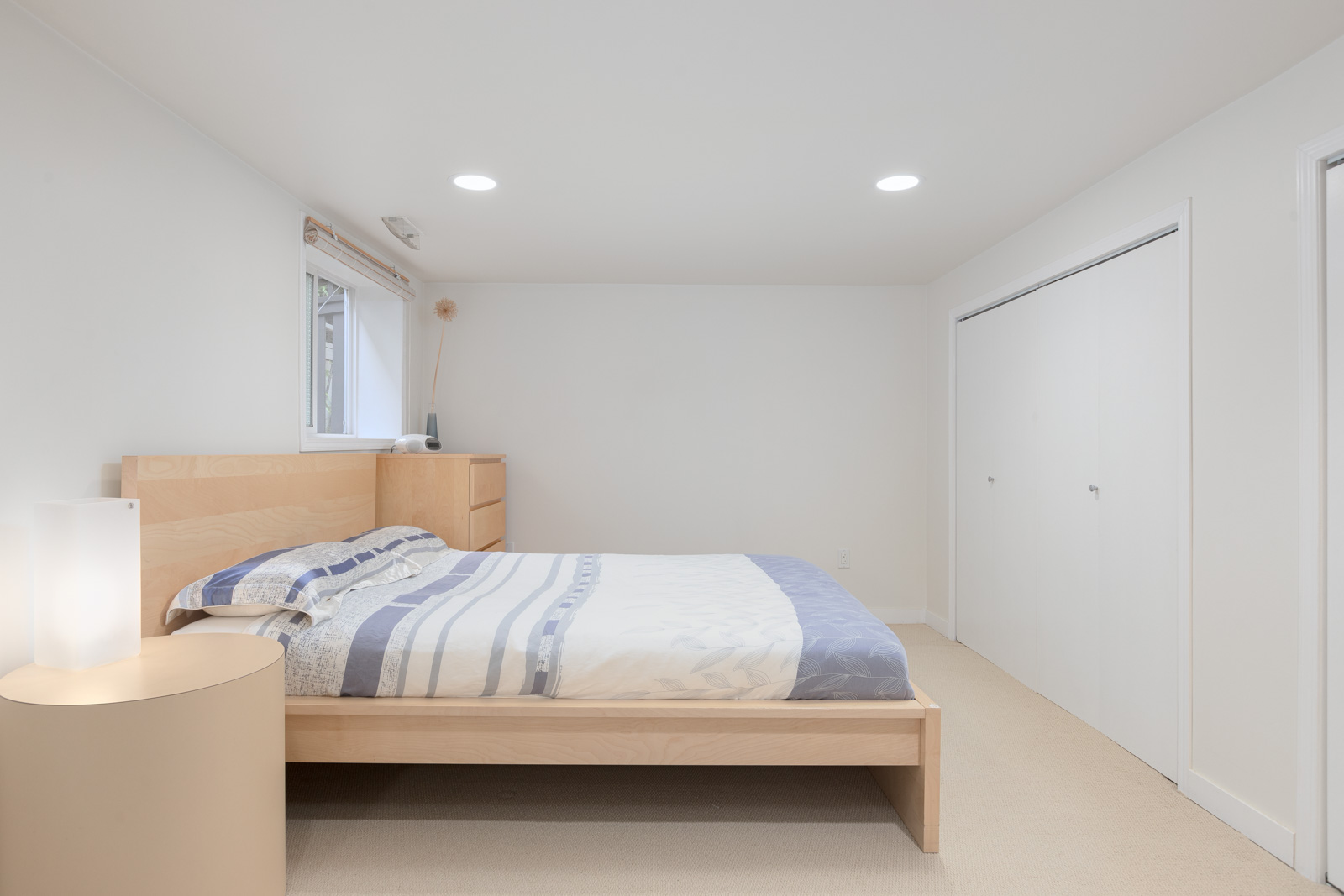 Minimalist bedroom with a wooden bed, striped bedding, a round nightstand with a lamp, built-in closet, dresser, and a window with natural light.