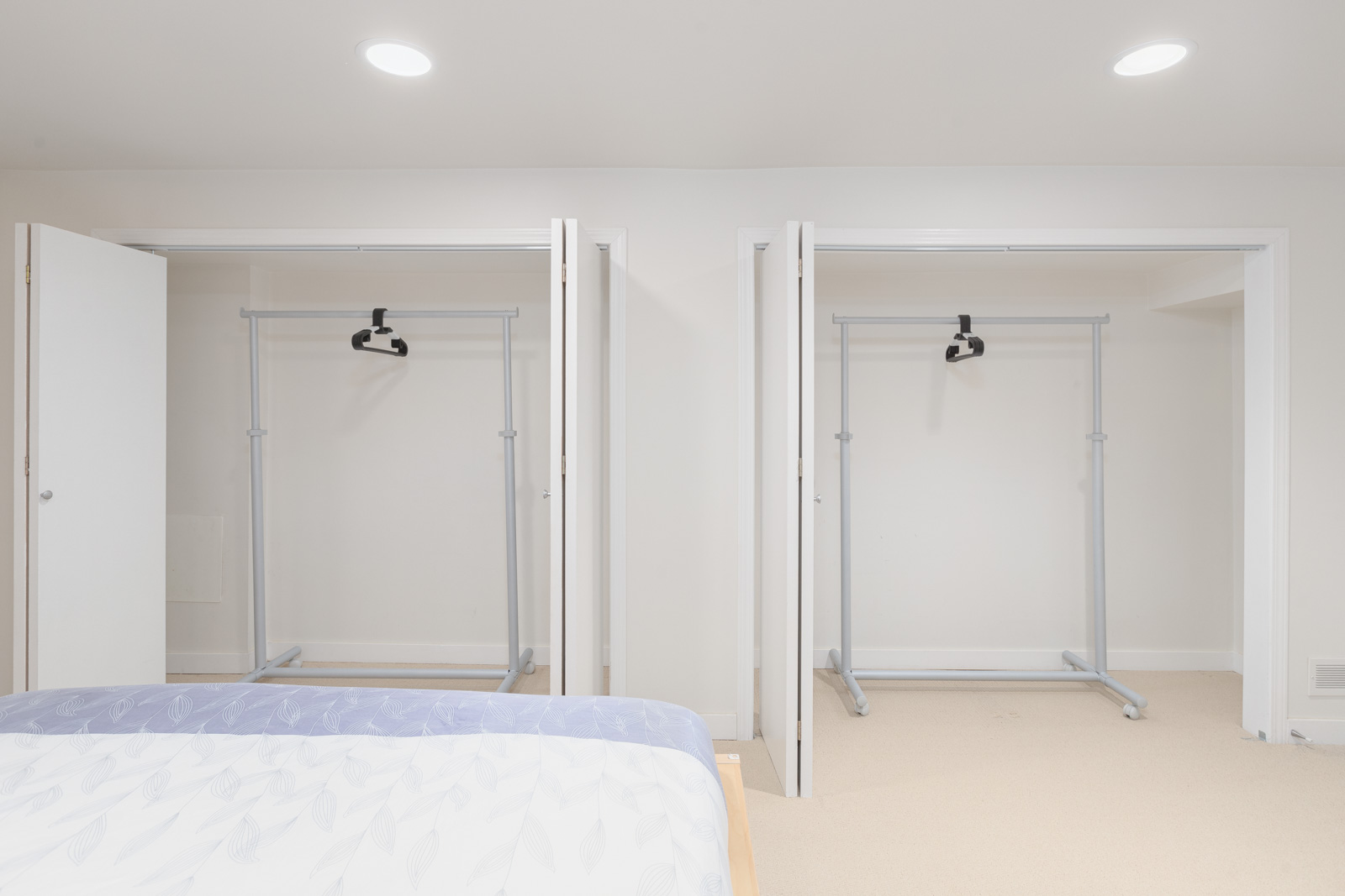 Two open closets in a bright, minimalist bedroom, each containing an empty metal clothing rack. Part of an unmade bed is visible in the foreground.