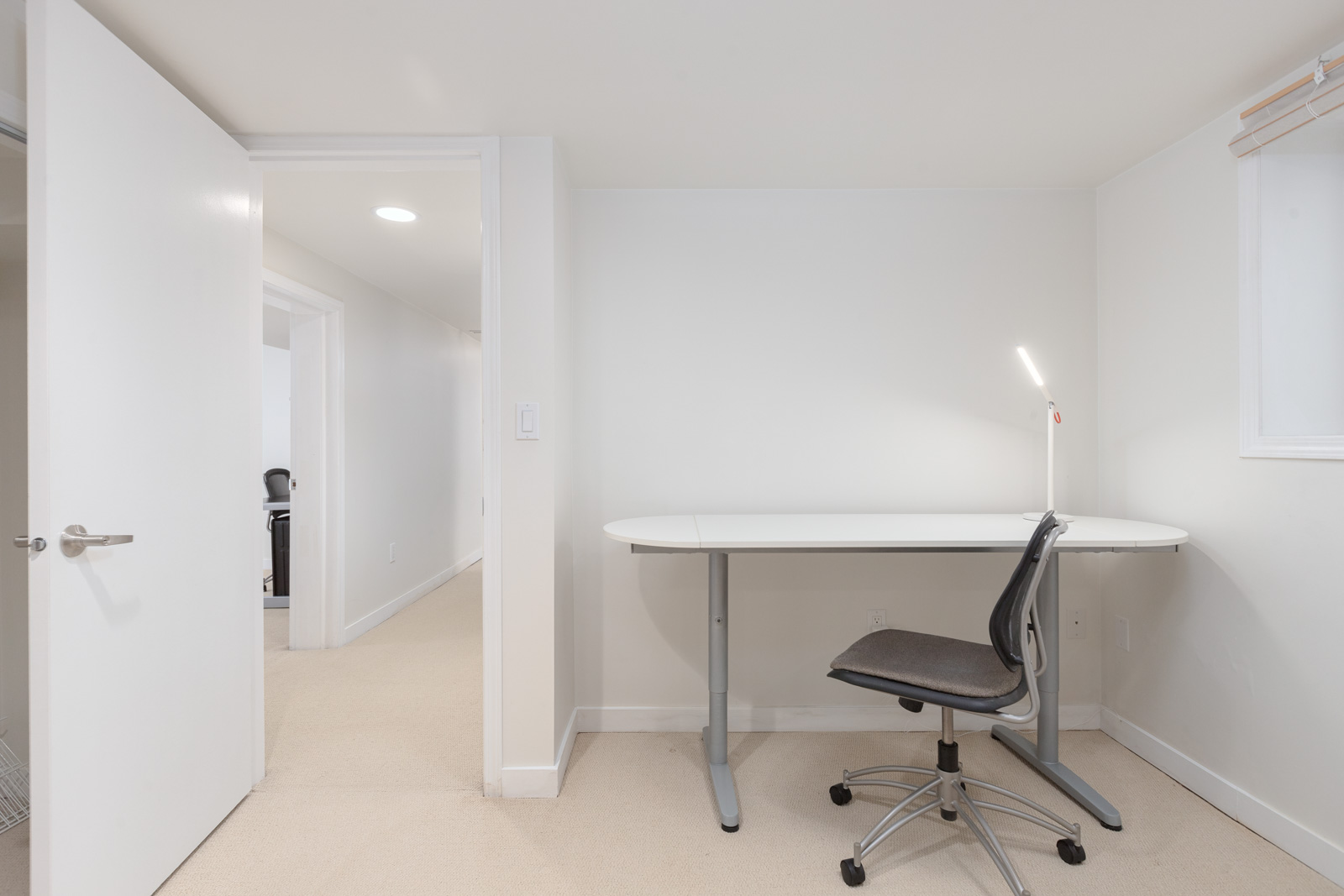 Minimalist office with a white desk, black office chair, white desk lamp, and open door leading to a hallway with beige carpet and white walls.