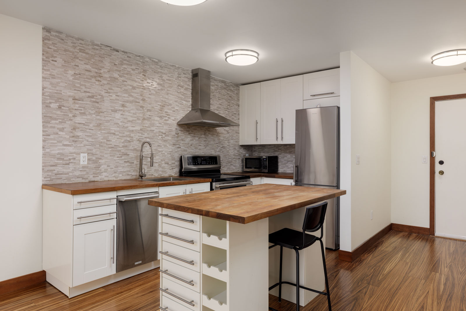 Modern kitchen with white cabinets, stainless steel appliances, wood countertops, a tile backsplash, and a small island with a single black chair.