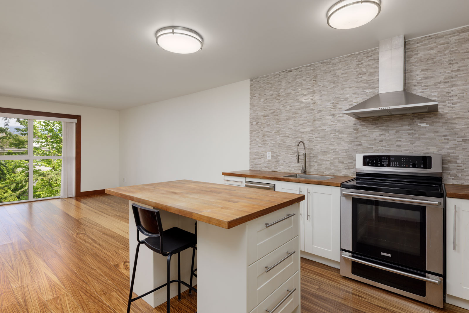 Modern kitchen with wood flooring, white cabinets, stainless steel appliances, a tile backsplash, and a small island with a single chair. Large window provides natural light.