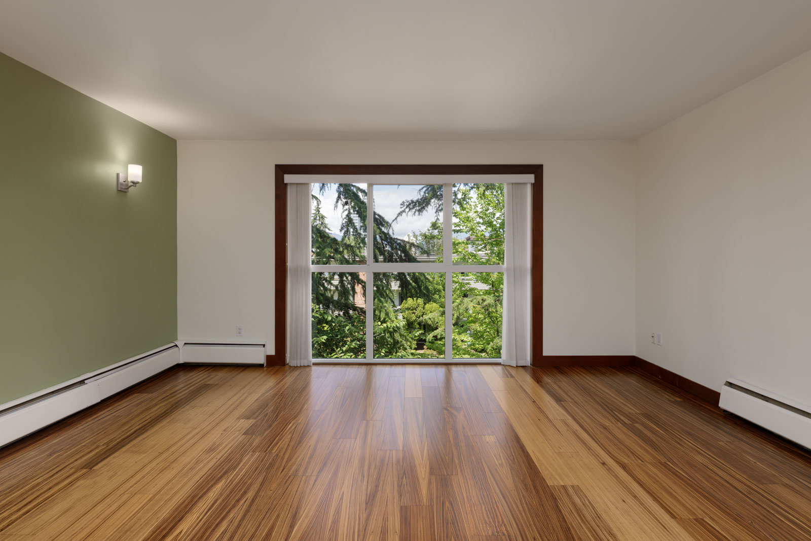 Empty room with wood flooring, green accent wall, white walls, large window with a view of trees, and wall-mounted light fixture.