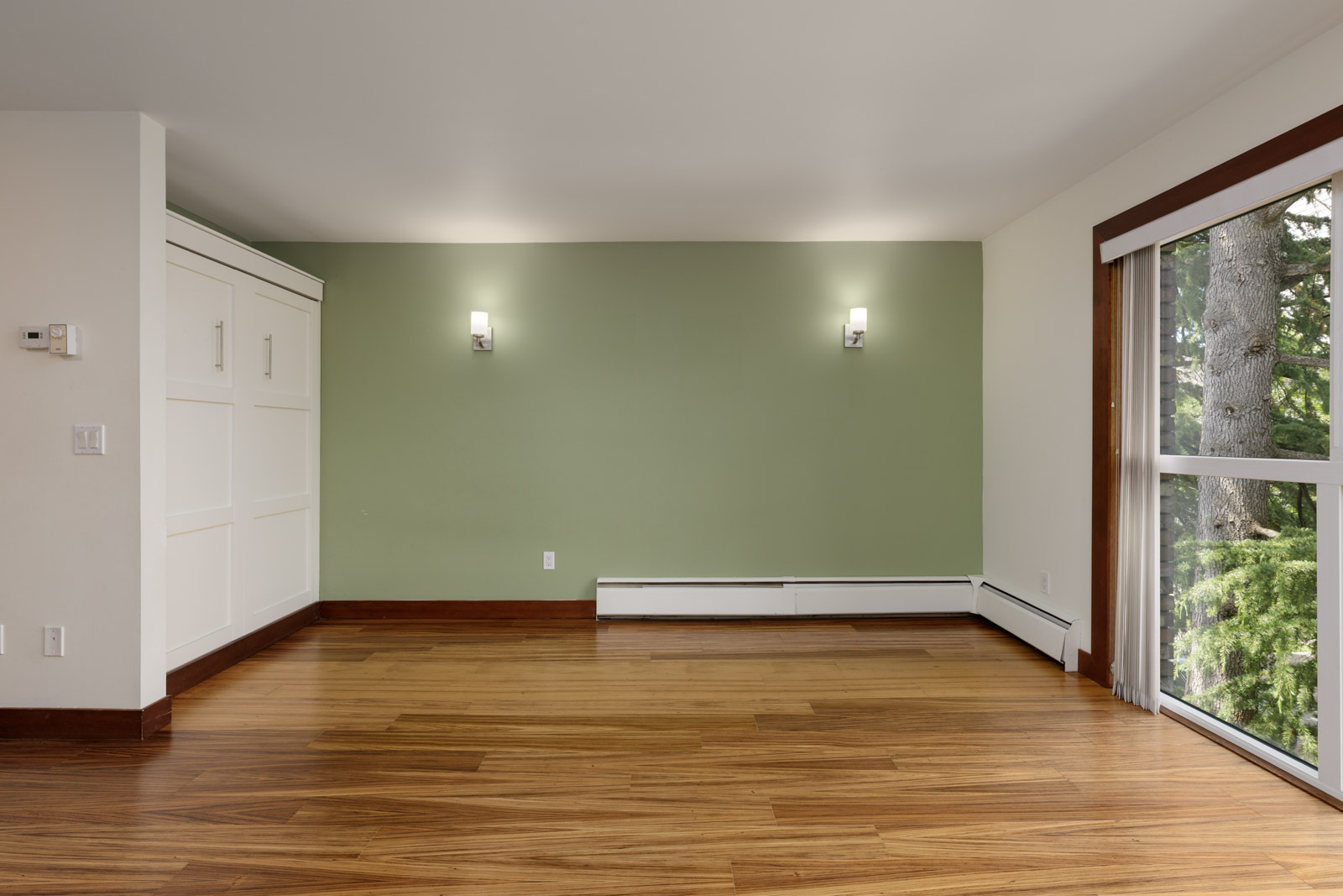 Empty room with wood floors, a built-in cabinet, green accent wall, two wall sconces, and a large window with a tree view.