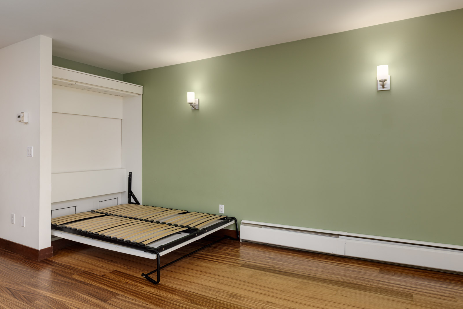A room with a green accent wall, wood flooring, two wall sconces, and an open Murphy bed frame without a mattress.