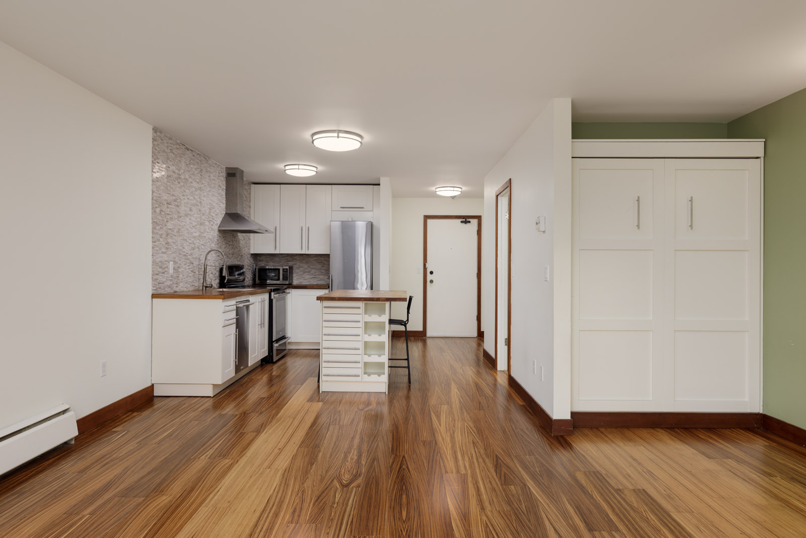 Modern apartment interior with wood flooring, compact kitchen with stainless steel appliances, and white cabinetry; fold-out Murphy bed on the right wall.