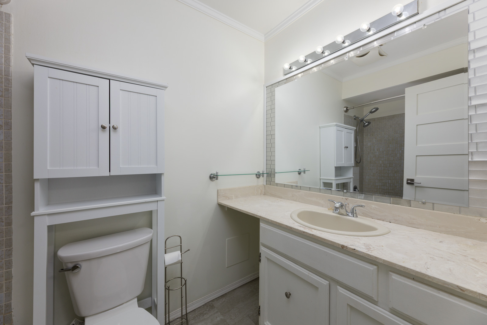 Bright bathroom with a large mirror, cream countertop with built-in sink, white cabinets, toilet, towel rack, and overhead vanity lights.