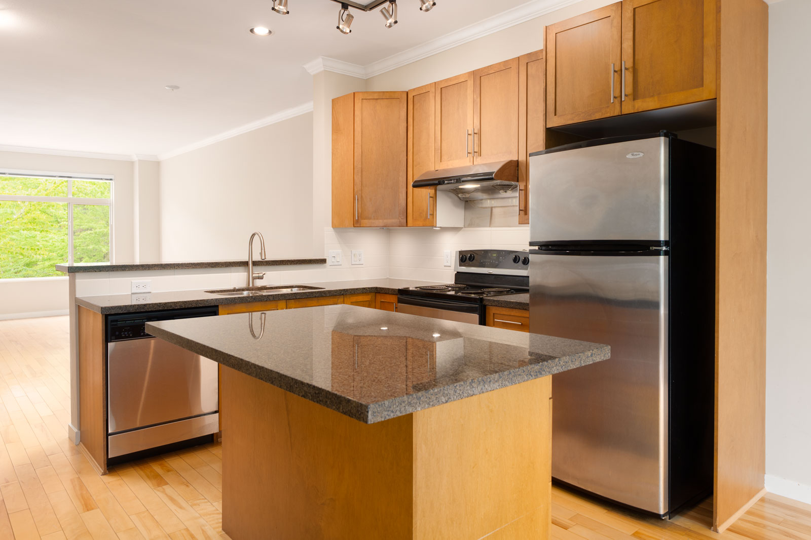 Modern kitchen with wooden cabinets, stainless steel appliances, a dishwasher, and a granite island countertop. Light wood flooring and a window in the adjoining room.