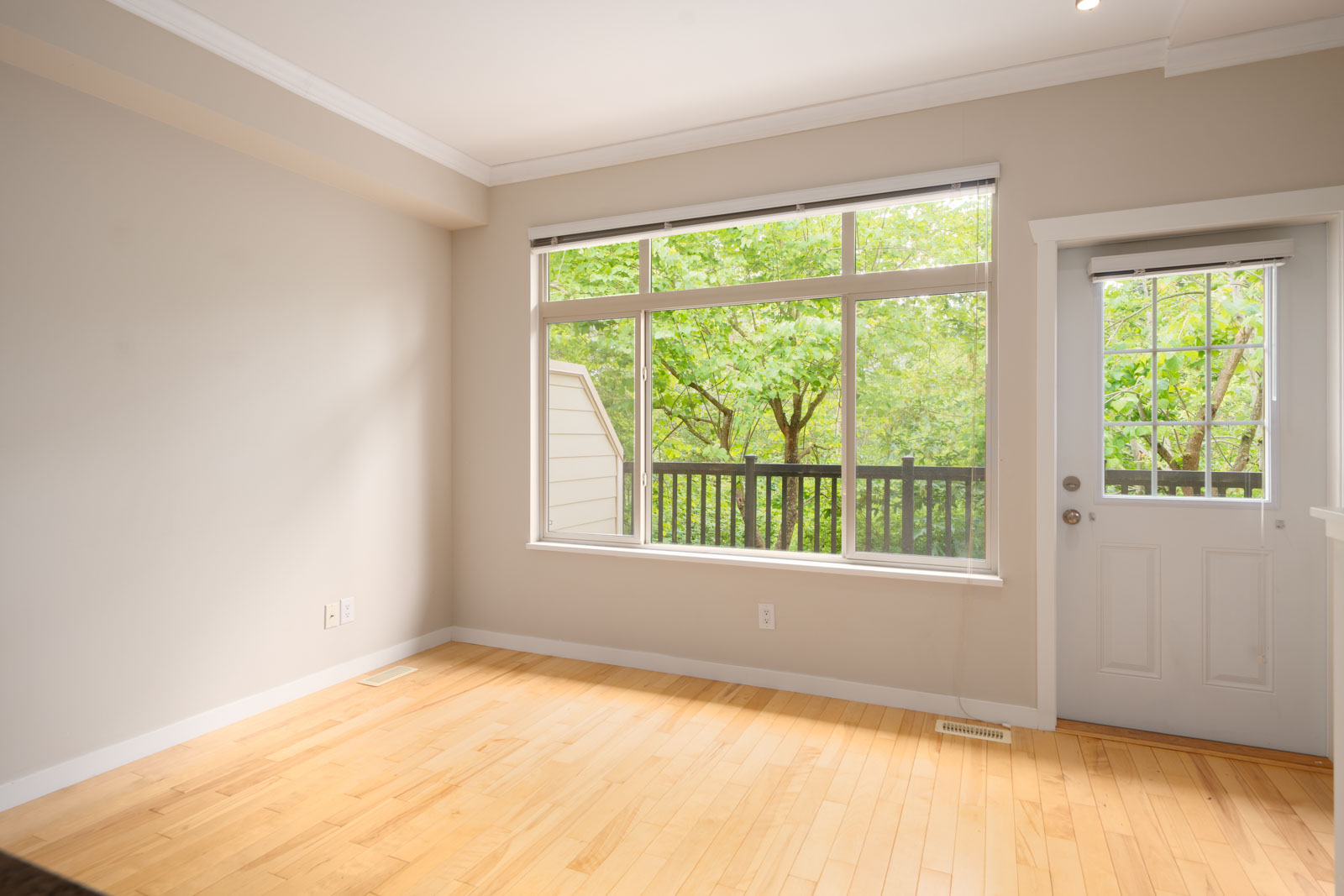 Empty room with light hardwood floor, beige walls, a large window, and a glass door that leads to a deck with trees visible outside.