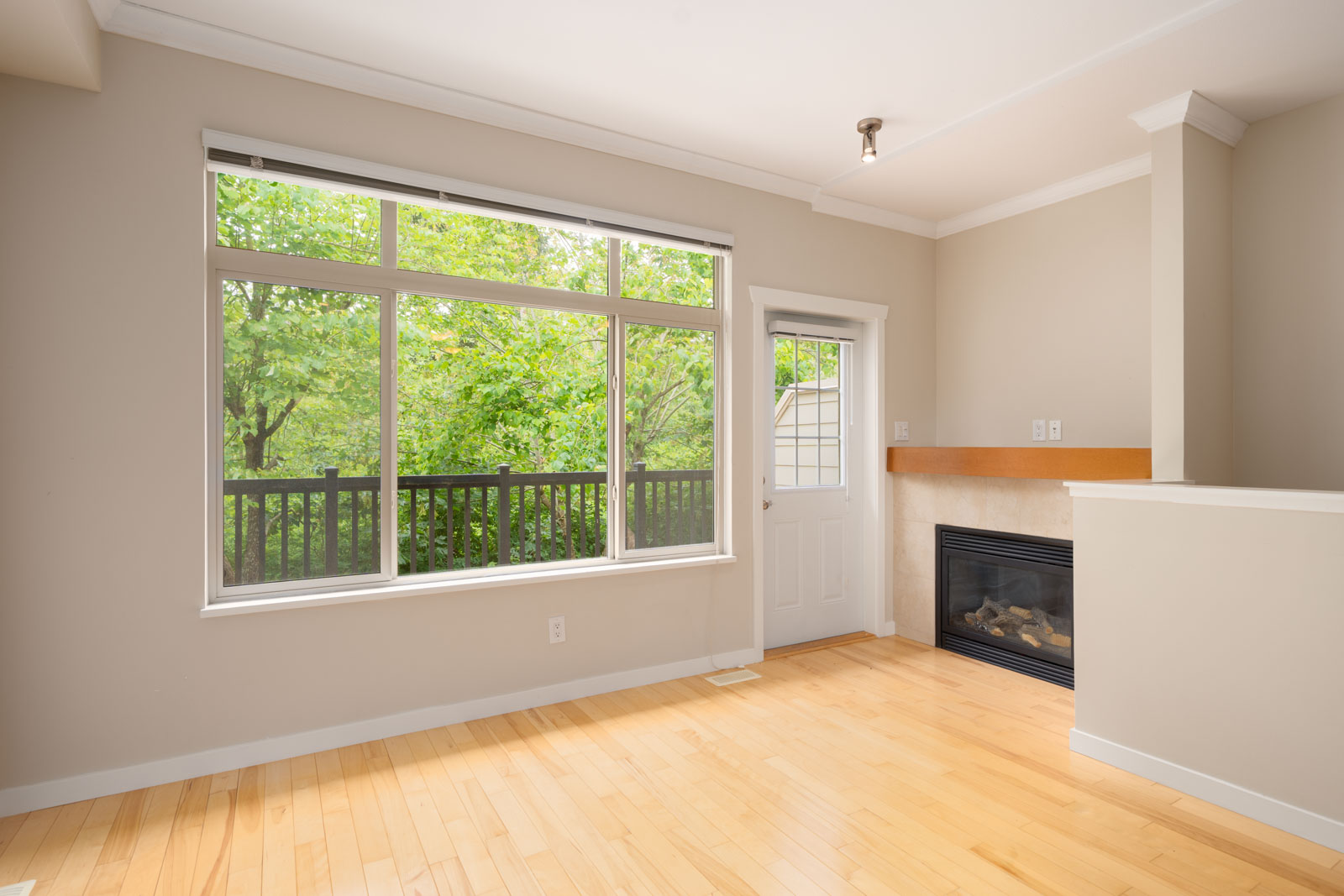 Bright, empty room with light wood floors, a large window, a glass door to a balcony, and a modern fireplace against a beige wall.