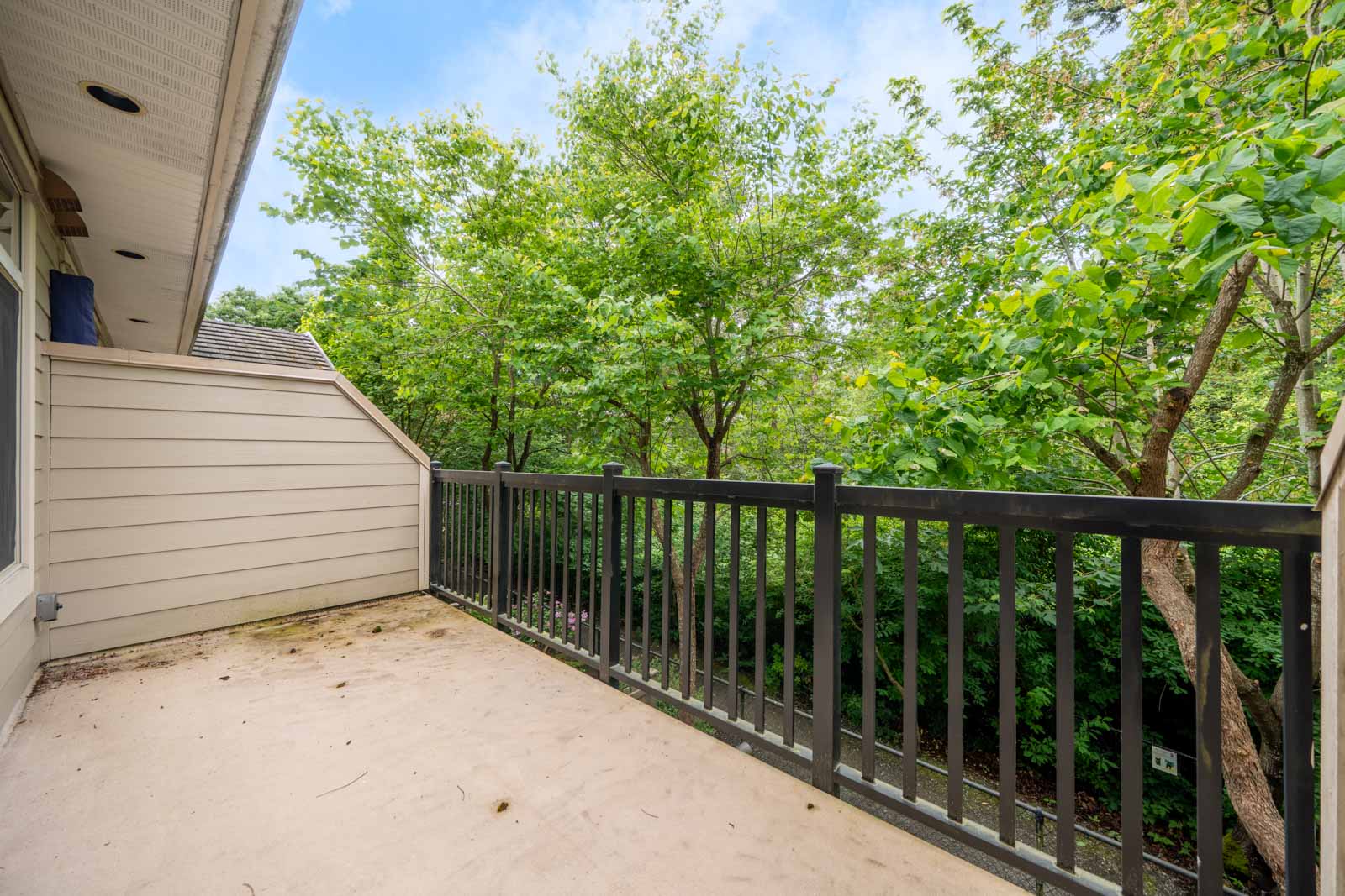 A small balcony with a black metal railing overlooks leafy green trees, attached to a beige-sided building under a partly cloudy sky.