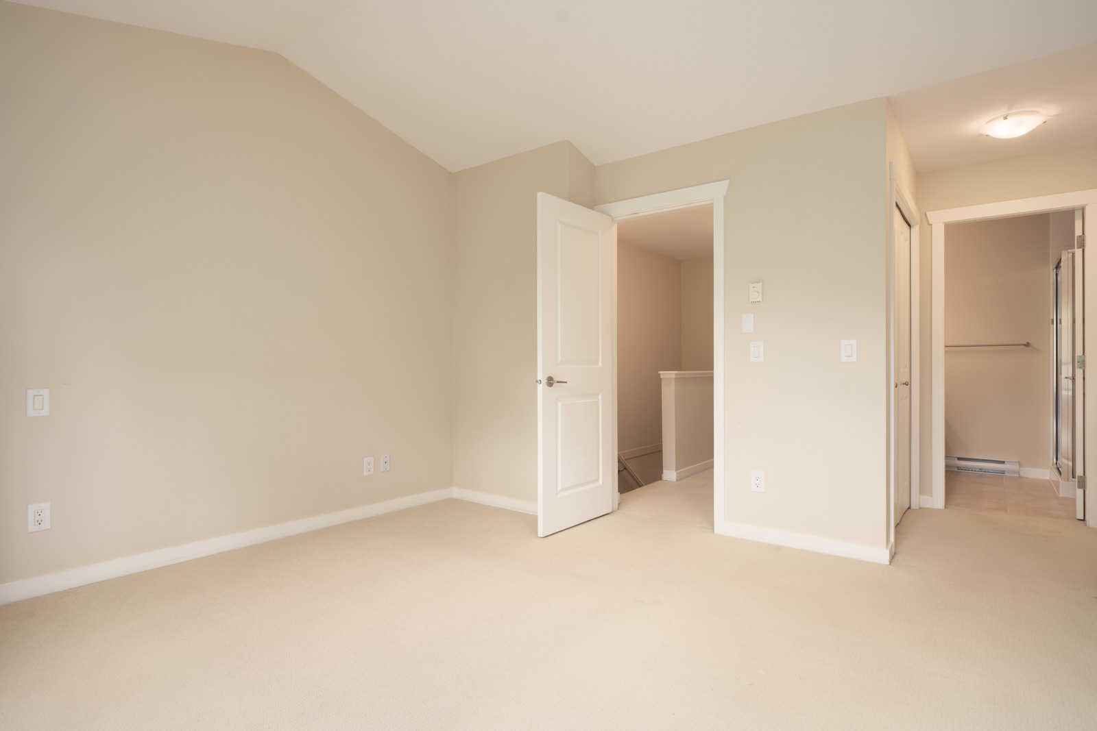Empty beige carpeted room with off-white walls, an open door leading to a hallway, and visible baseboards and light switches.