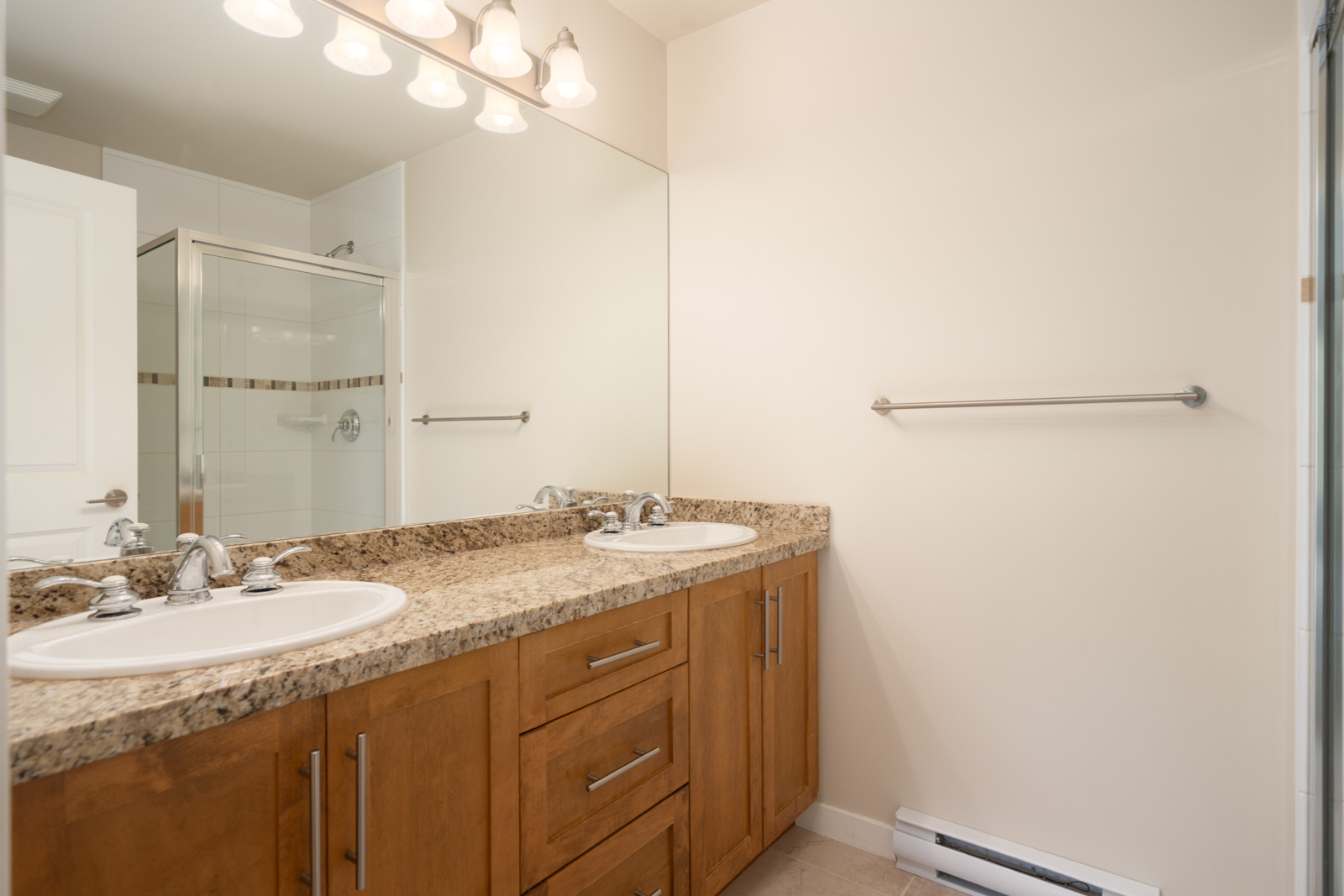 Double sink bathroom vanity with granite countertop, wooden cabinets, large wall mirror, and a towel bar on a beige wall; shower enclosure visible in the background.