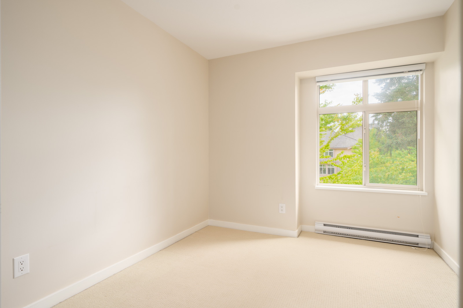 Empty room with beige walls, carpeted floor, a window with a view of greenery, and a baseboard heater beneath the window.