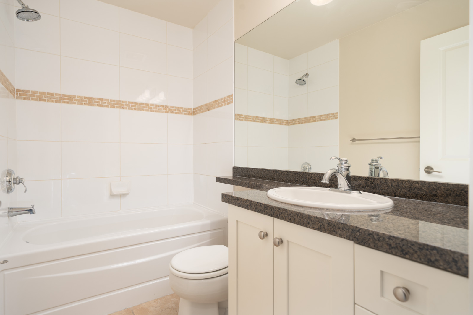 Bright bathroom with a bathtub, toilet, and sink on a dark countertop, large mirror above, and white tiled walls with a decorative accent strip.