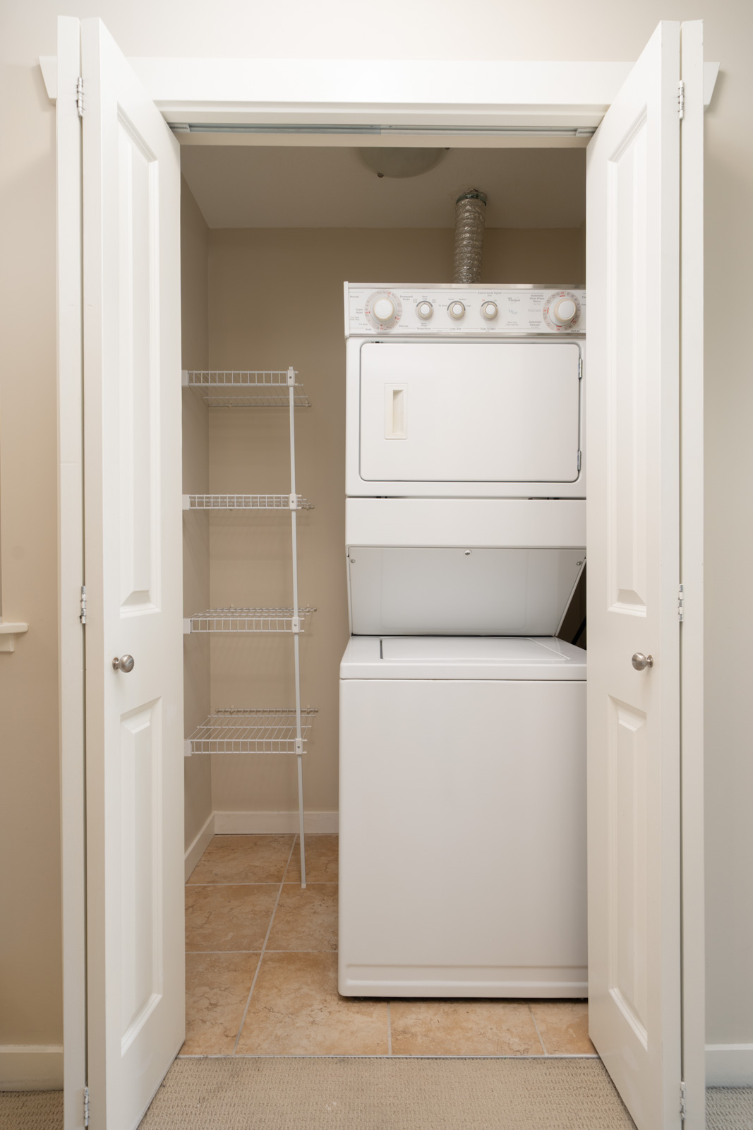 A small laundry closet with white folding doors, containing a stacked washer and dryer unit and wire shelving on a tiled floor.