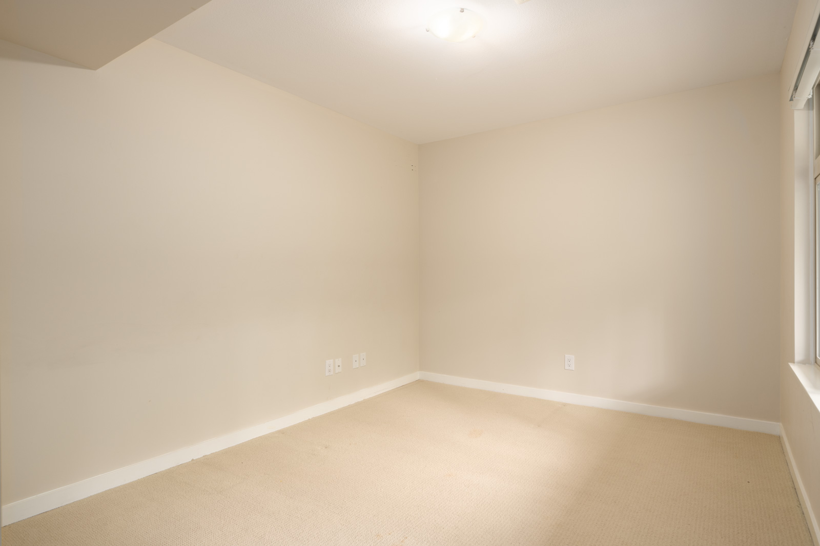 Empty room with beige carpet, off-white walls, ceiling light, and electrical outlets on the left wall. Natural light enters from a window on the right.