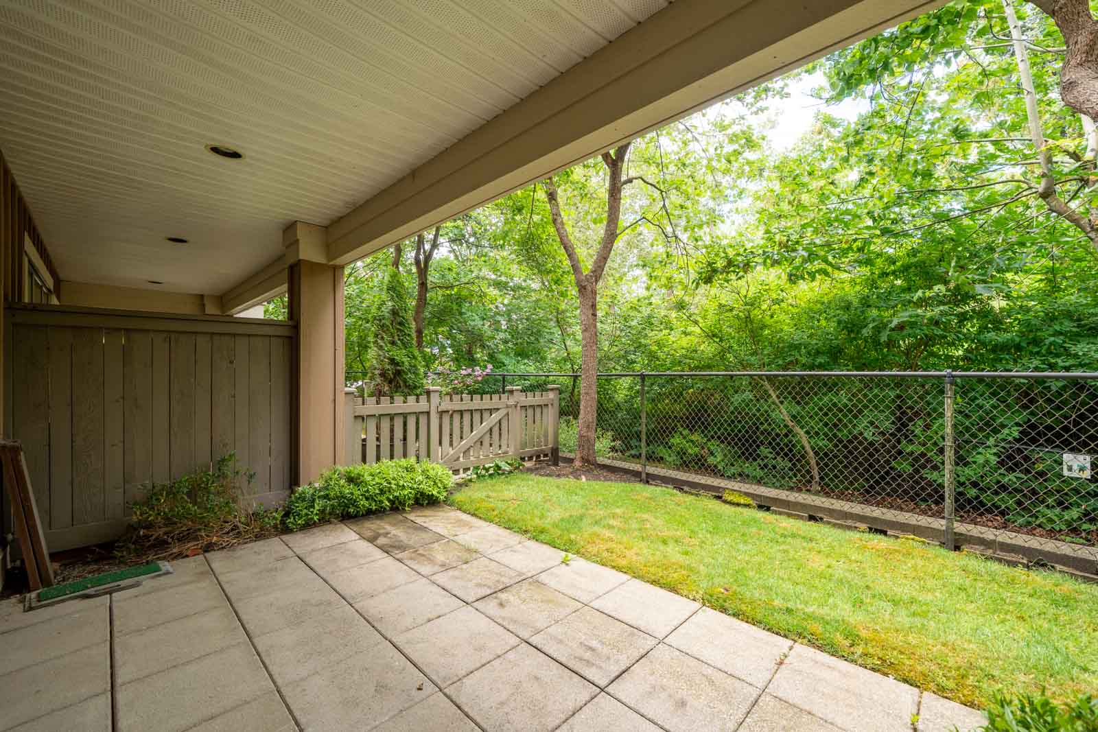Covered patio with tiled floor, small grassy area, and fence, overlooking a view of trees and greenery in a backyard.