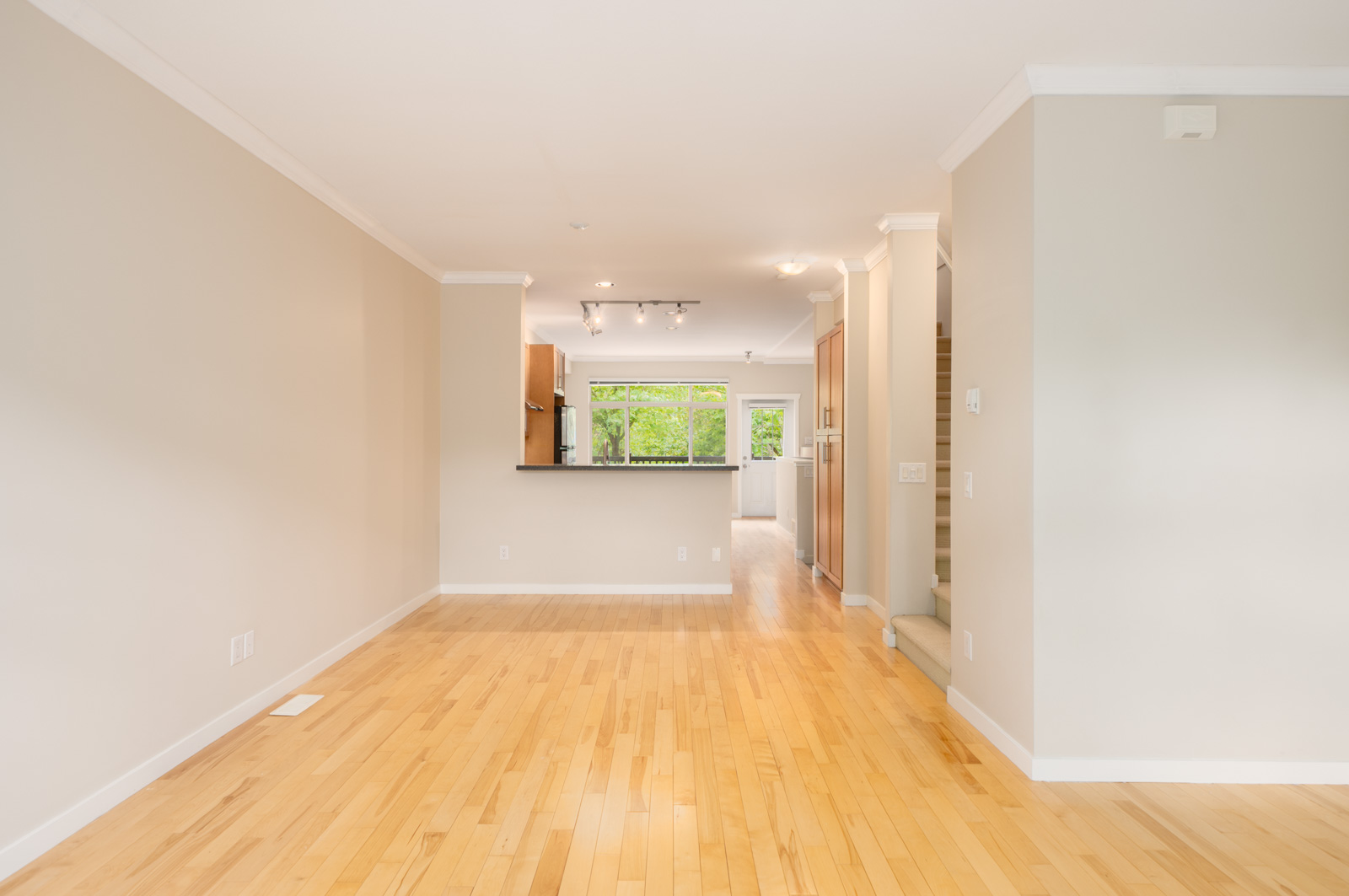 Empty room with light wood floors, beige walls, and white trim, leading to an open kitchen and large windows with greenery visible outside.