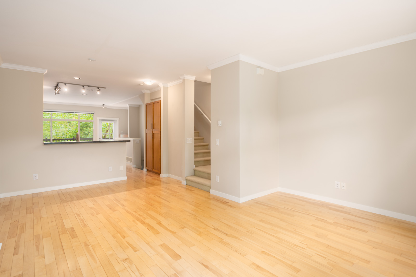 Bright empty living space with light wood floors, beige walls, open kitchen with bar counter, recessed lights, and stairway leading upstairs.
