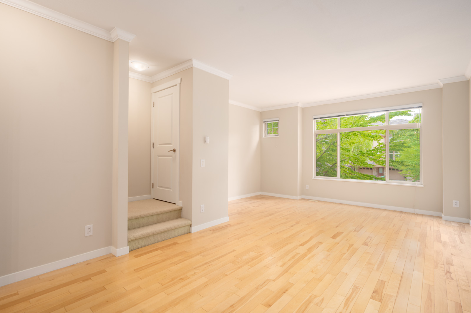 Empty living room with light wood floors, beige walls, a large window with green trees outside, and a small staircase leading to a closed door.