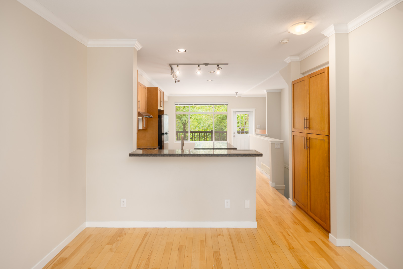 Modern kitchen with light wood floors, beige walls, wood cabinets, a granite countertop, overhead lighting, and a view of a bright living area with large windows and greenery outside.