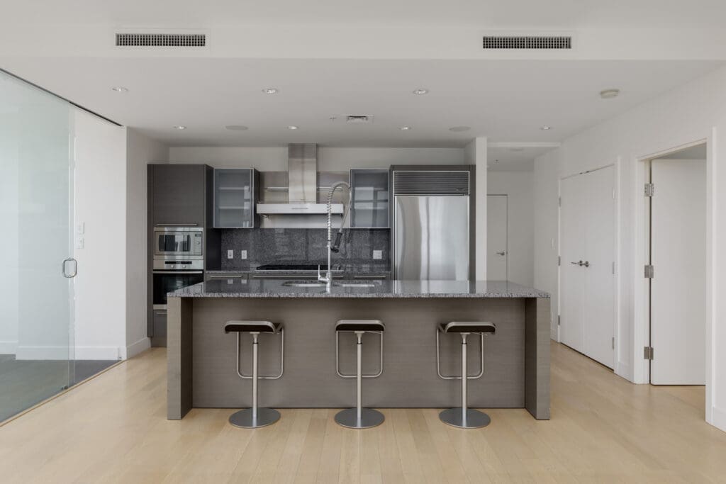 Modern kitchen with an island, three bar stools, stainless steel appliances, grey cabinets, and light wood flooring.