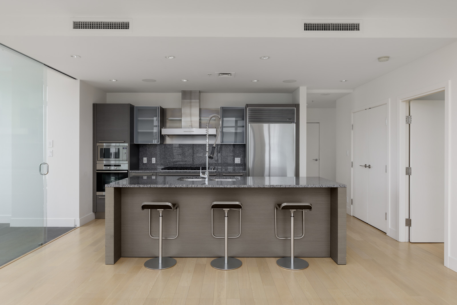 Modern kitchen with an island, three bar stools, stainless steel appliances, grey cabinets, and light wood flooring.