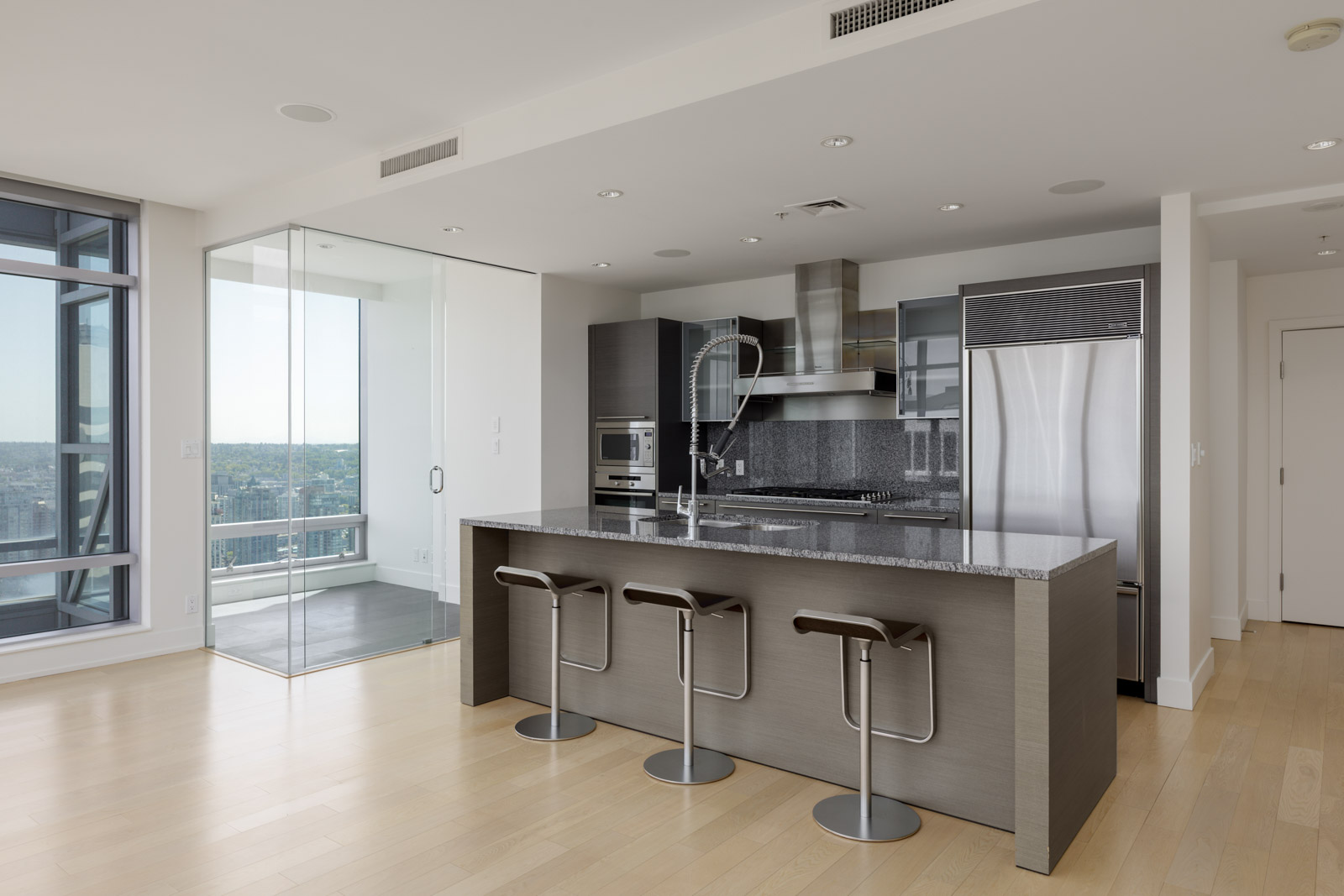 Modern kitchen with dark cabinets, stainless steel appliances, and a large island with three barstools; glass doors open to a balcony with city views.