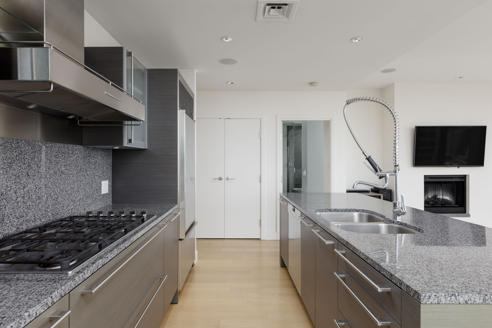Modern kitchen with gray cabinets, granite countertops, gas stove, island with sink, wall-mounted TV, and light wood flooring.