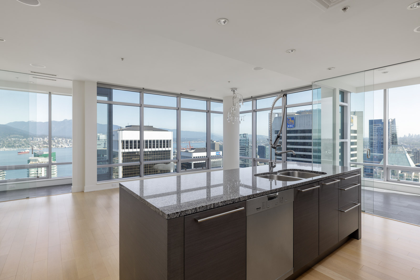 Modern high-rise kitchen with granite island, stainless steel appliances, and floor-to-ceiling windows overlooking a city skyline and waterfront.